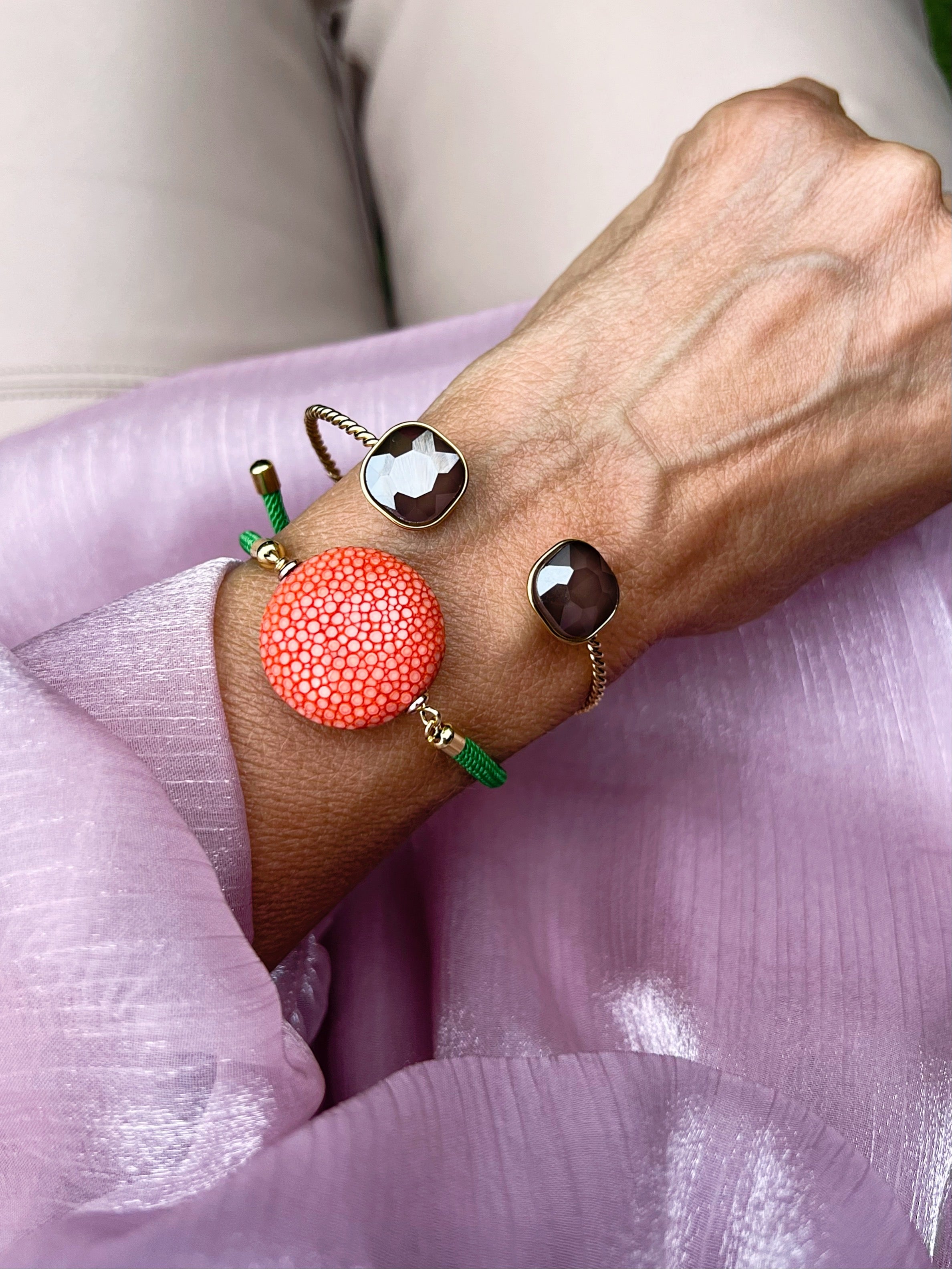 Ein grünes Armband mit oranger Perle dazu ein goldener Armreif mit Glasstein in Taupe. An einem Handgelenk auf rosa Stoff fotografiert.