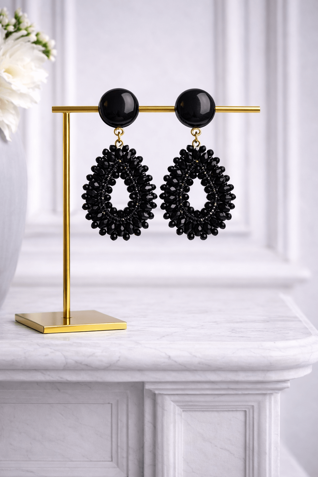 Schwarze tropfenförmige Ohrringe mit Garnbasis und dicht angeordneten schwarzen Glasperlen, befestigt an runden schwarzen Ohrsteckern, präsentiert auf goldenem Schmuckständer.