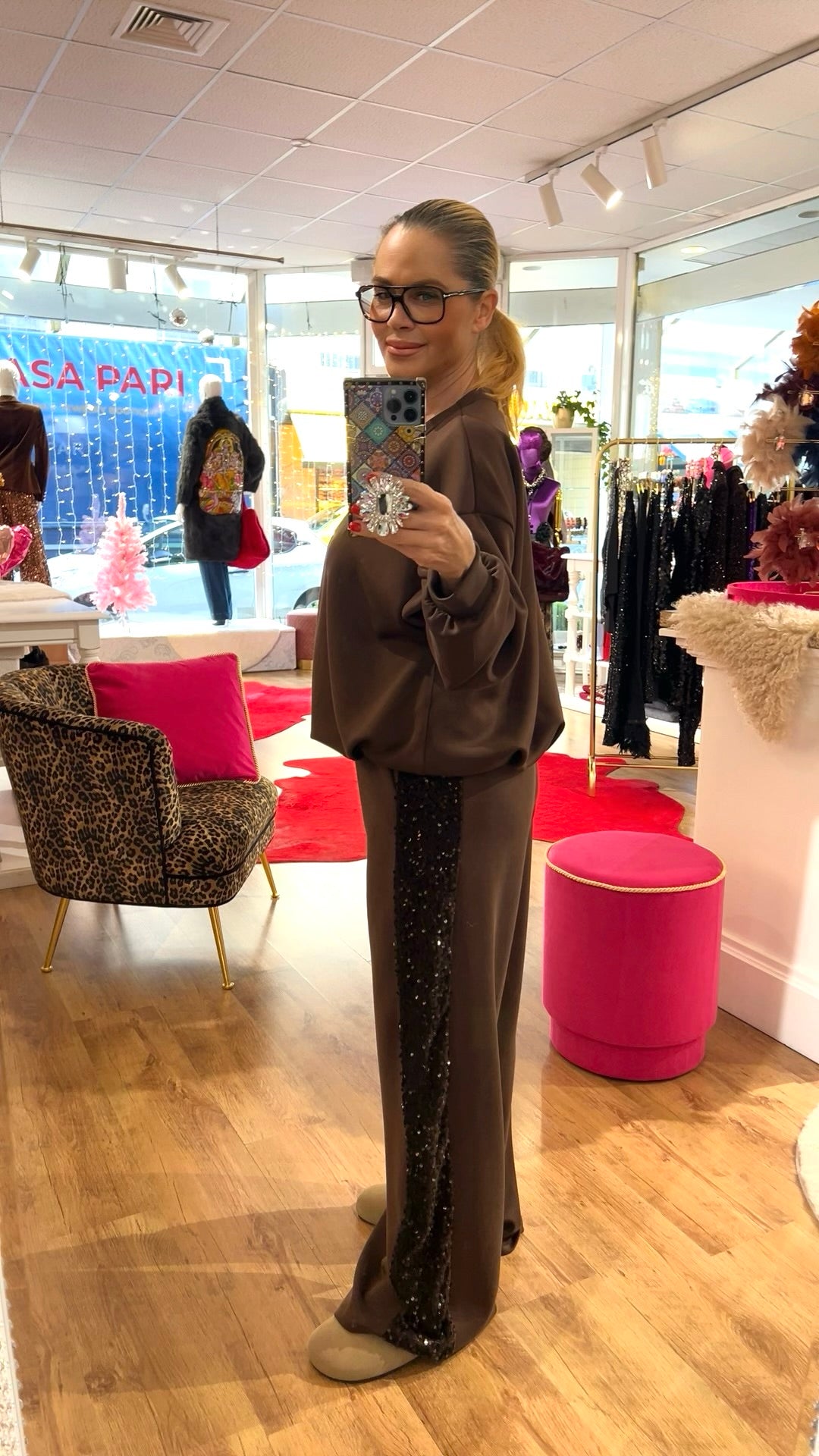 Seitenansicht einer Person in einem braunen Lounge-Set mit lockerem Oberteil und Paillettenstreifen an der Hose, aufgenommen in einer Boutique.
