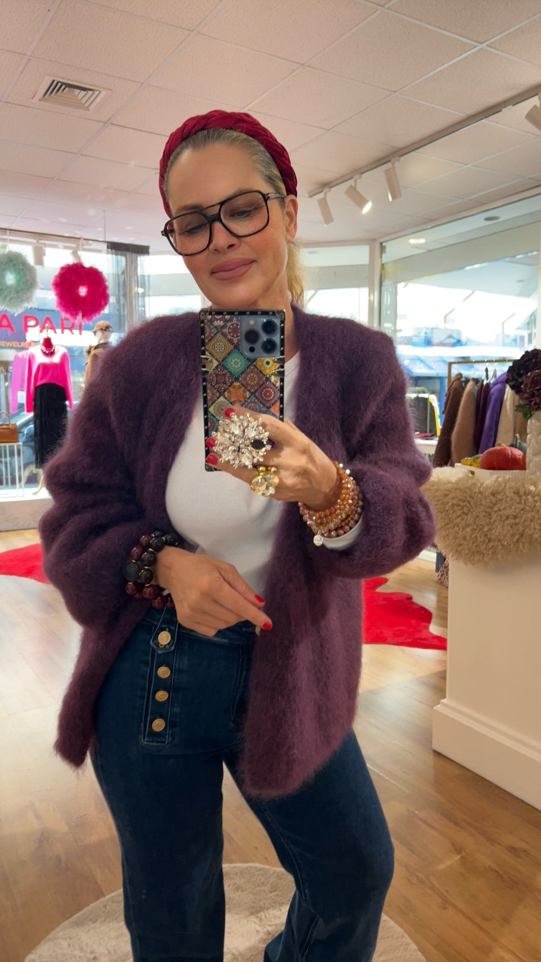 Frau trägt flauschige Strickjacke in Aubergine, kombiniert mit weißem Shirt und dunkelblauer Jeans mit Knopfleiste. Im Hintergrund bunte Mode und rote Akzente.