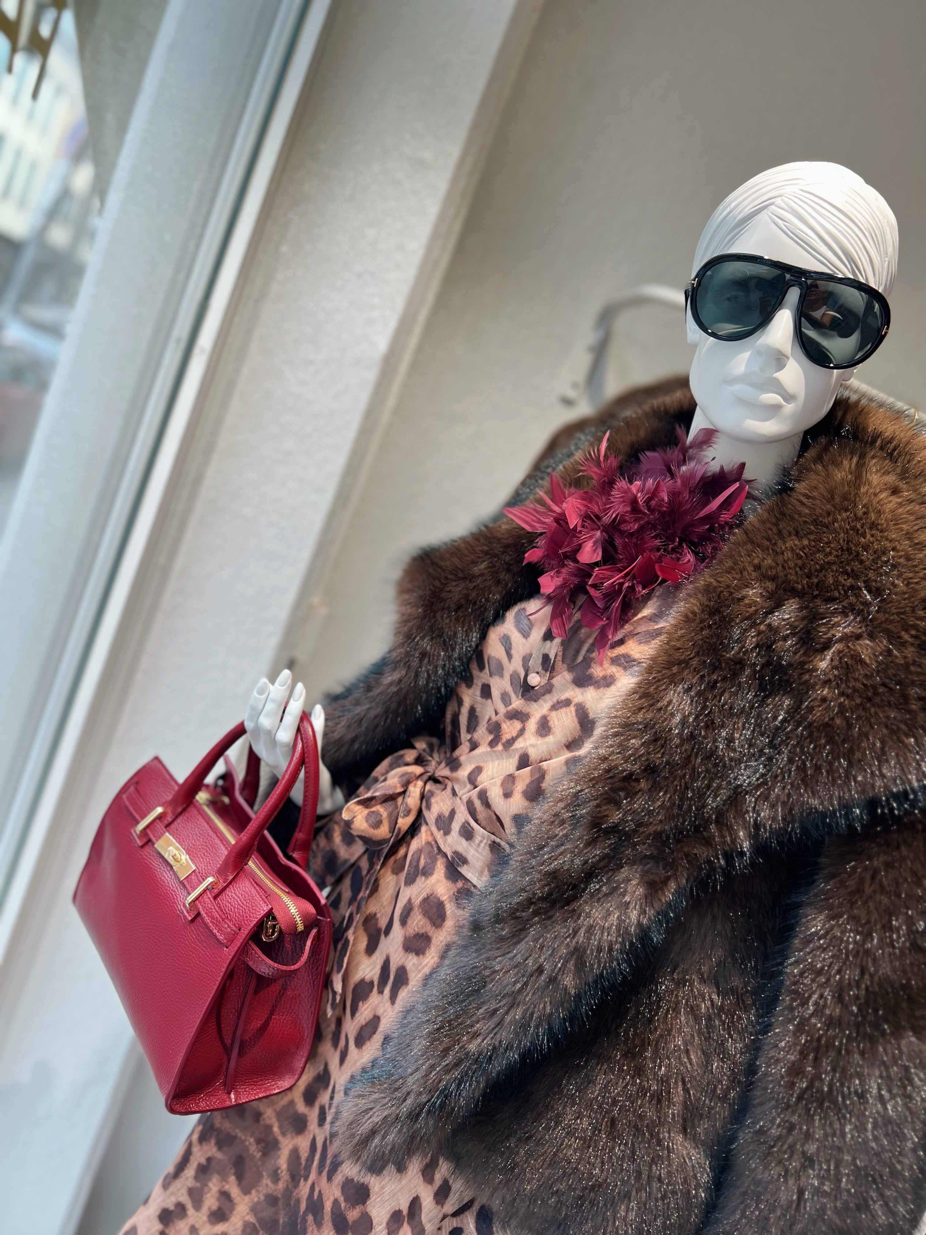 Schaufensterpuppe trägt braune Pelzjacke ein Leo Kleid und eine Handtasche in Rot, mit einer Sonnebrille. Am Kragen eine rote Federbrosche.