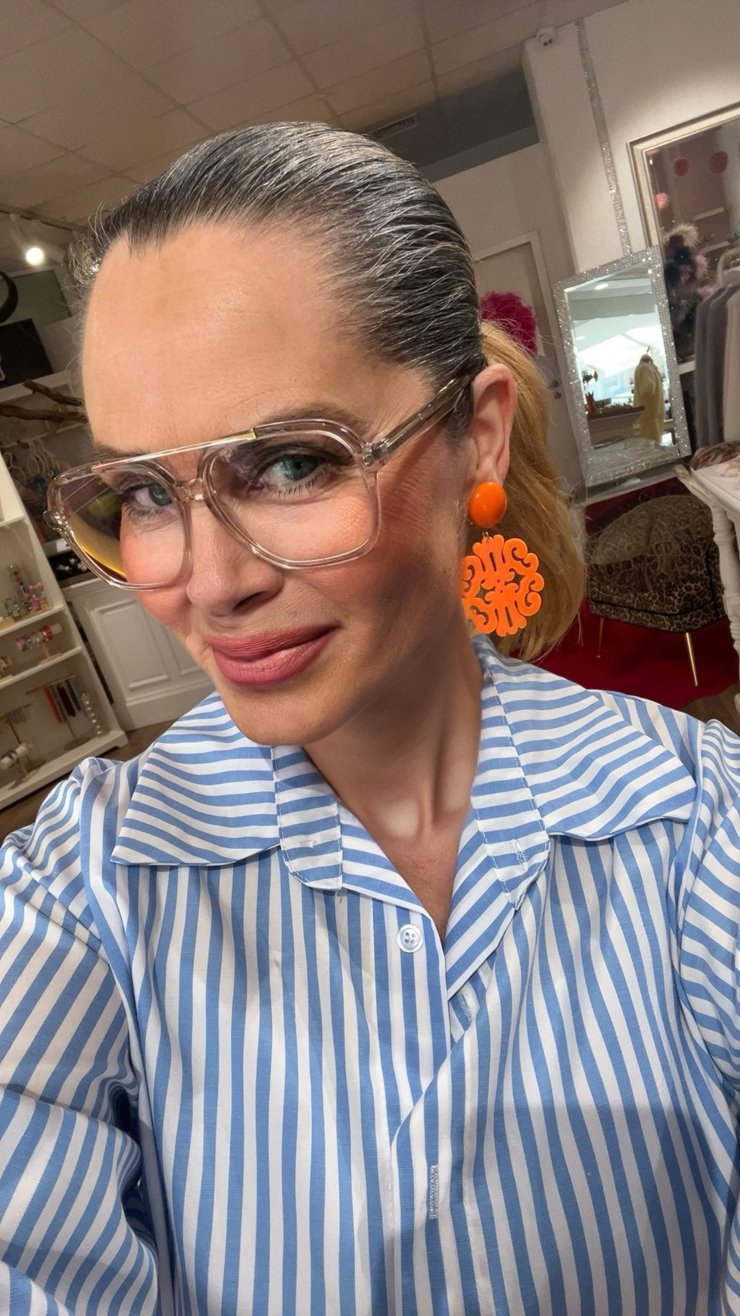 Frau mit zurückgebundenem Haar trägt eine blau-weiß gestreifte Bluse, transparente Brille und große orangefarbene Statement-Ohrringe, aufgenommen in einer Boutique mit Regalen und Spiegel im Hintergrund.
