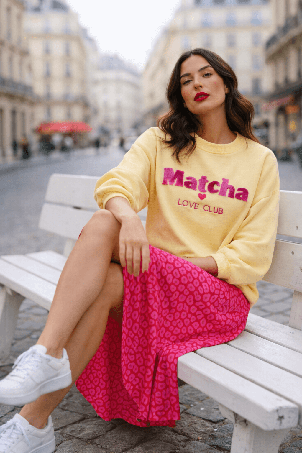 Frau sitzt auf einer weißen Holzbank in einer Pariser Innenstadtstraße. Sie trägt einen pinken Midi-Rock mit Leo-Muster, einen gelben Sweater mit der Aufschrift „Matcha Love Club“ und weiße Sneaker.