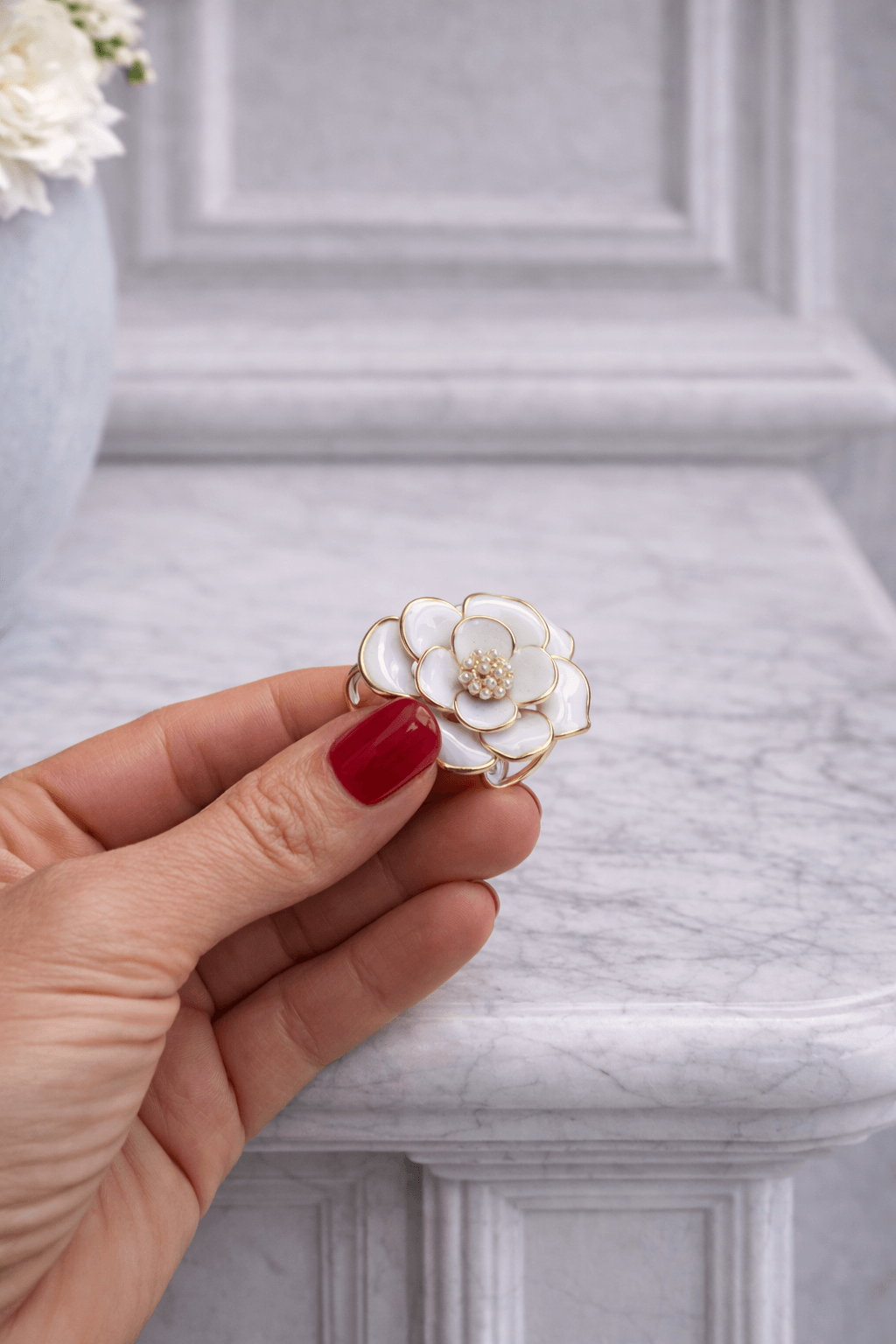 Hand mit rot lackierten Fingernägeln hält einen goldfarbenen Schalring mit weißer Blütenform und Perlendetail vor hellem Marmorhintergrund.