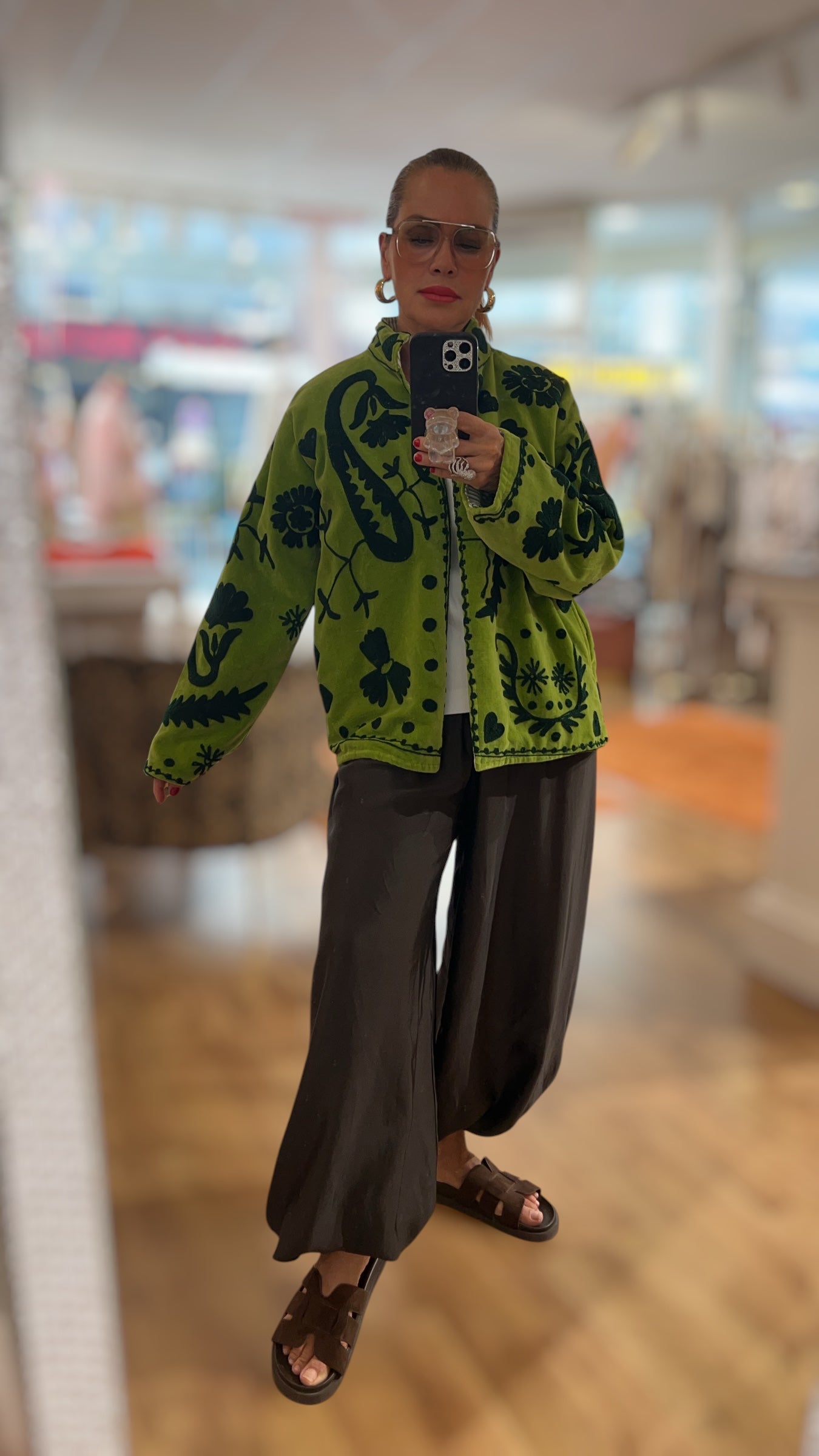 Frau trägt eine grüne gemusterte Jacke mit dunklem floralen Ornamentprint, Stehkragen und Knopfleiste. Kombiniert mit einer weiten schwarzen Hose und flachen Sandalen, fotografiert in einer hellen Boutique