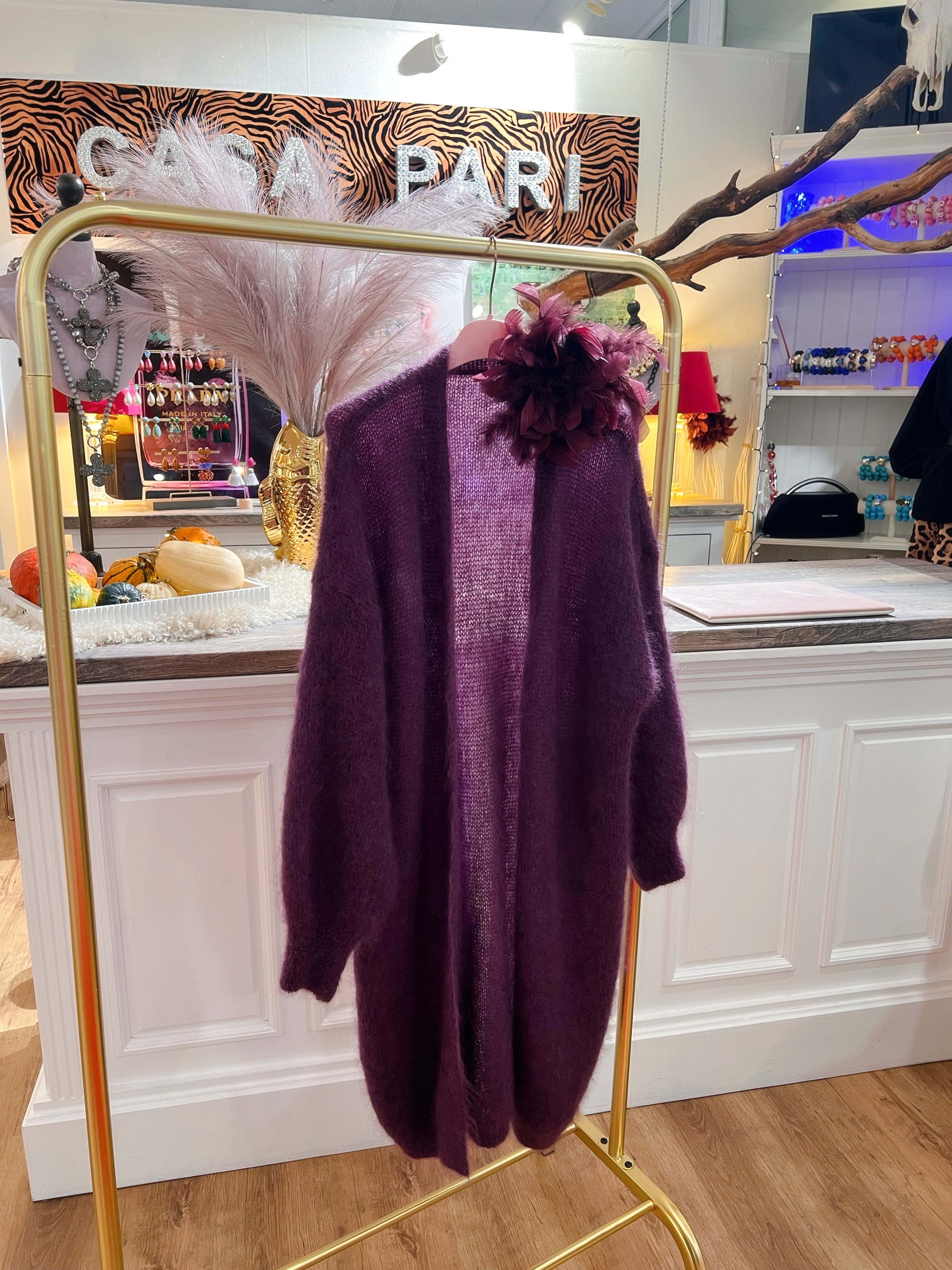 Ein weicher Kid- Mohair Cardigan hängt auf rosafarbenen Bügel an einer goldenen Kleiderstange in einer Boutique. Ein einem V der andere in dunklem Violett. Mit Blume am Kragen.