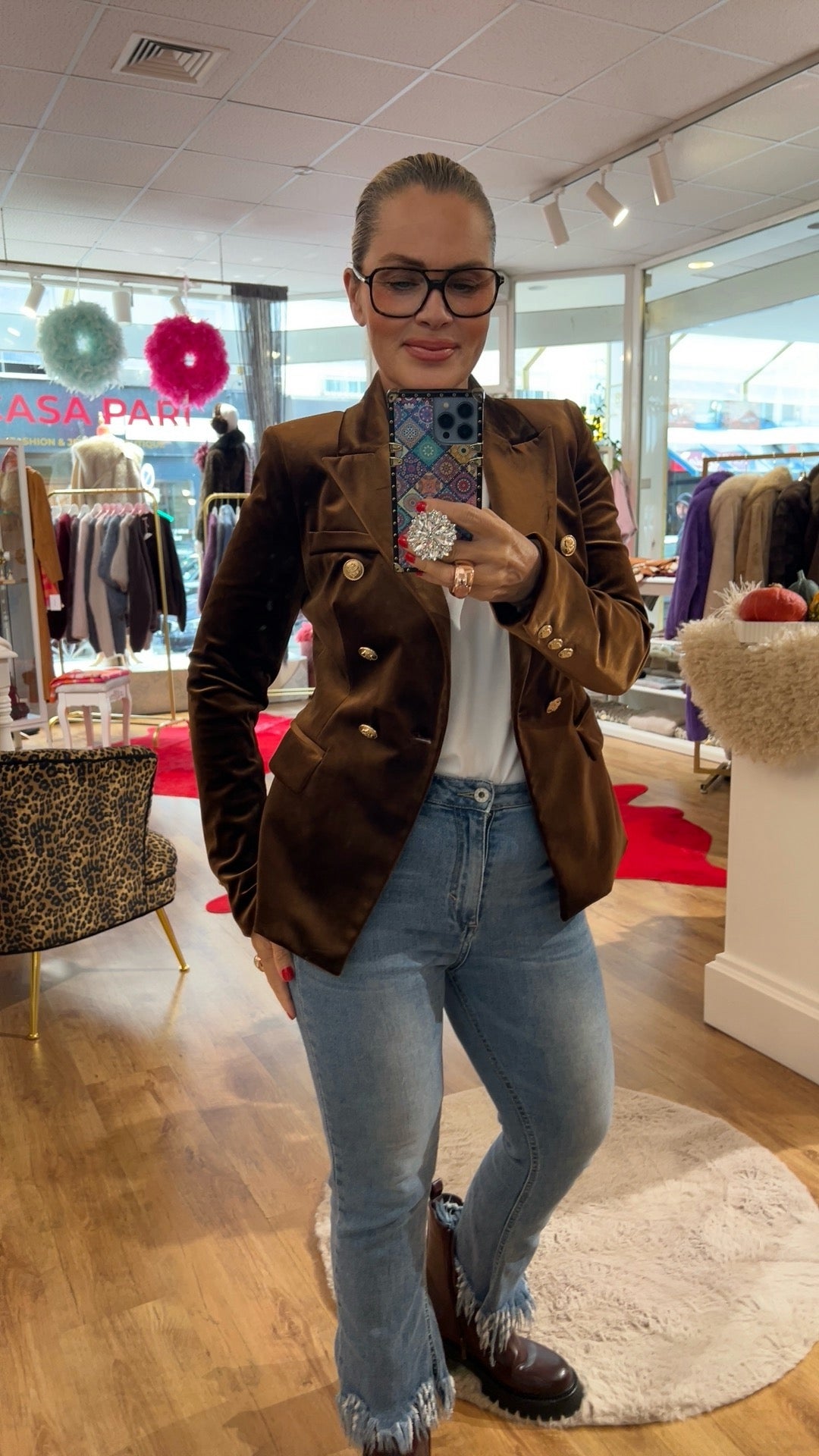 Frau trägt einen dunkel braunen Samtblazer mit goldenen Knöpfen. Dazu eine Jeans mit braunen Stiefeln. Sie hält ein Handy in der Hand. Fotografiert in einer Boutique.