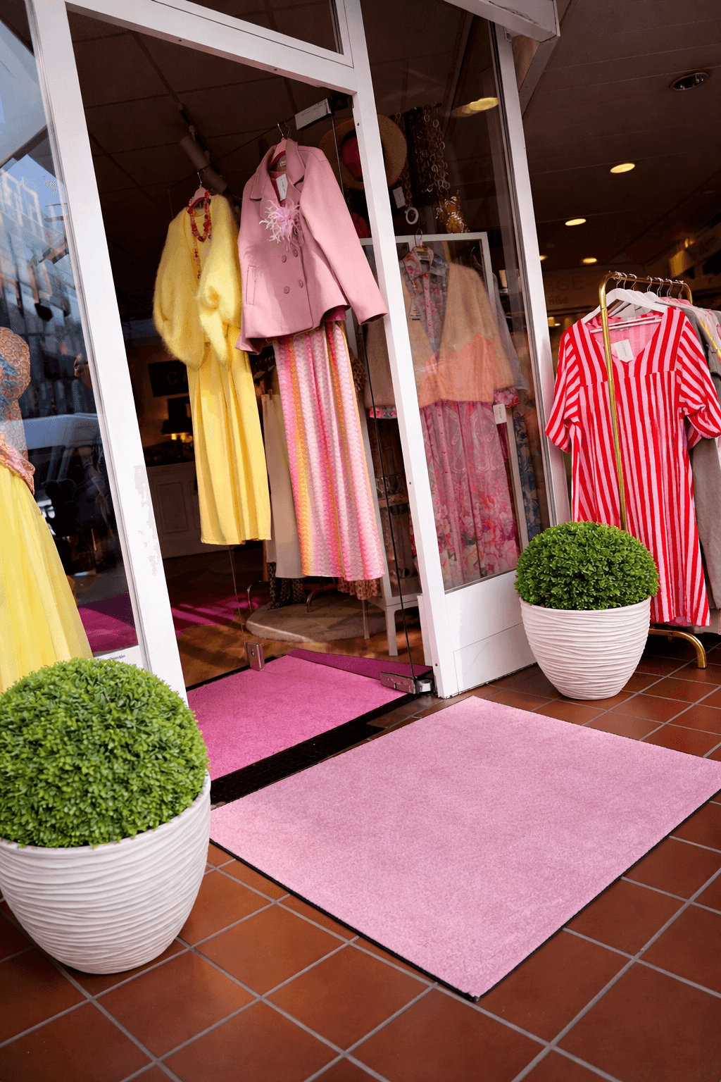 Eingang eines Modegeschäftes mit Kleiderständer, Schaufensterpuppen und pinker Fußmatte.