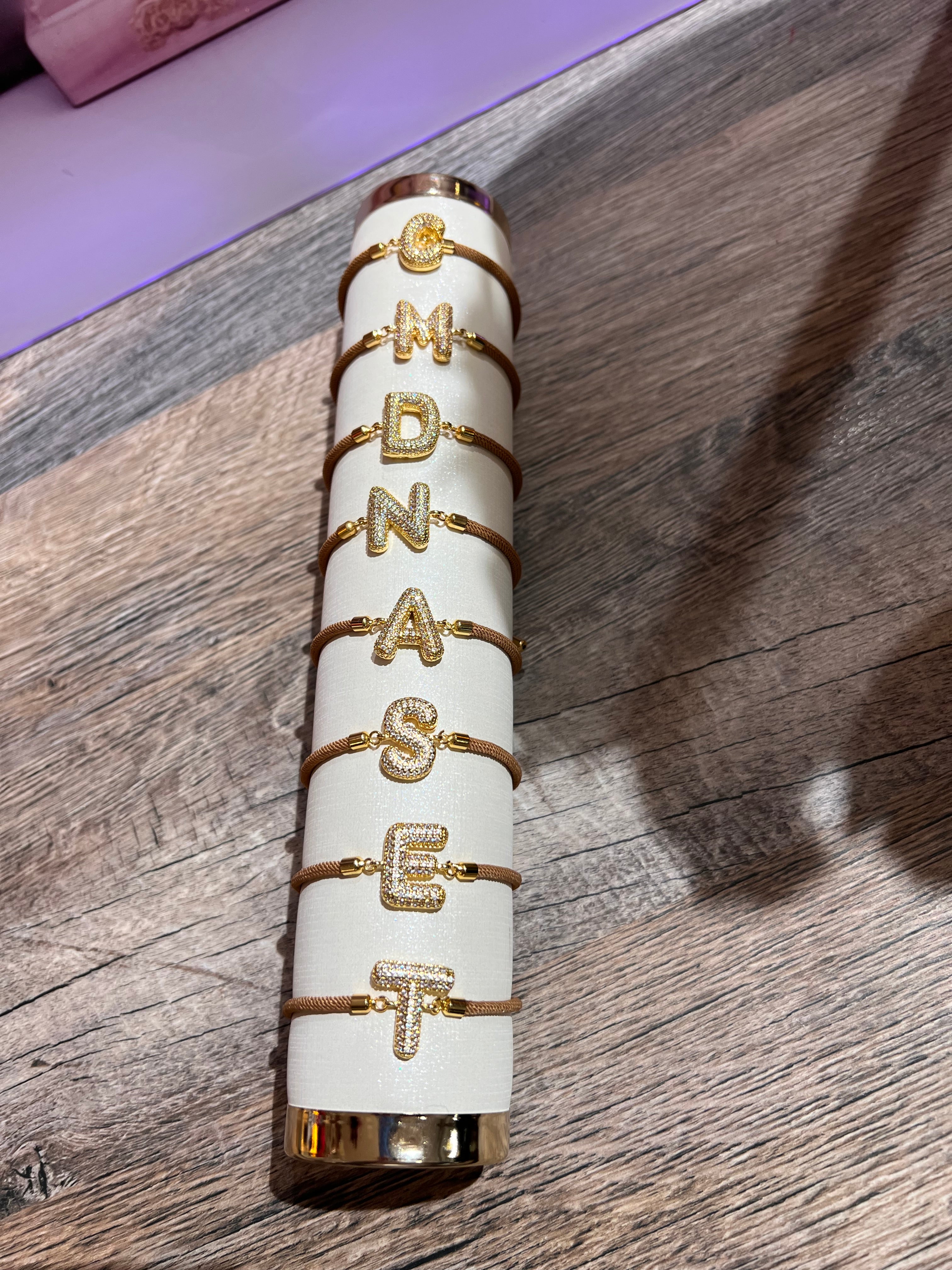 Mehrere goldfarbene Armbänder mit glitzernden Buchstaben Elementen C, M, D, N, A, S, E, und T auf einem cremefarbenen Präsentationsständer, liegend auf einer Holzoberfläche.