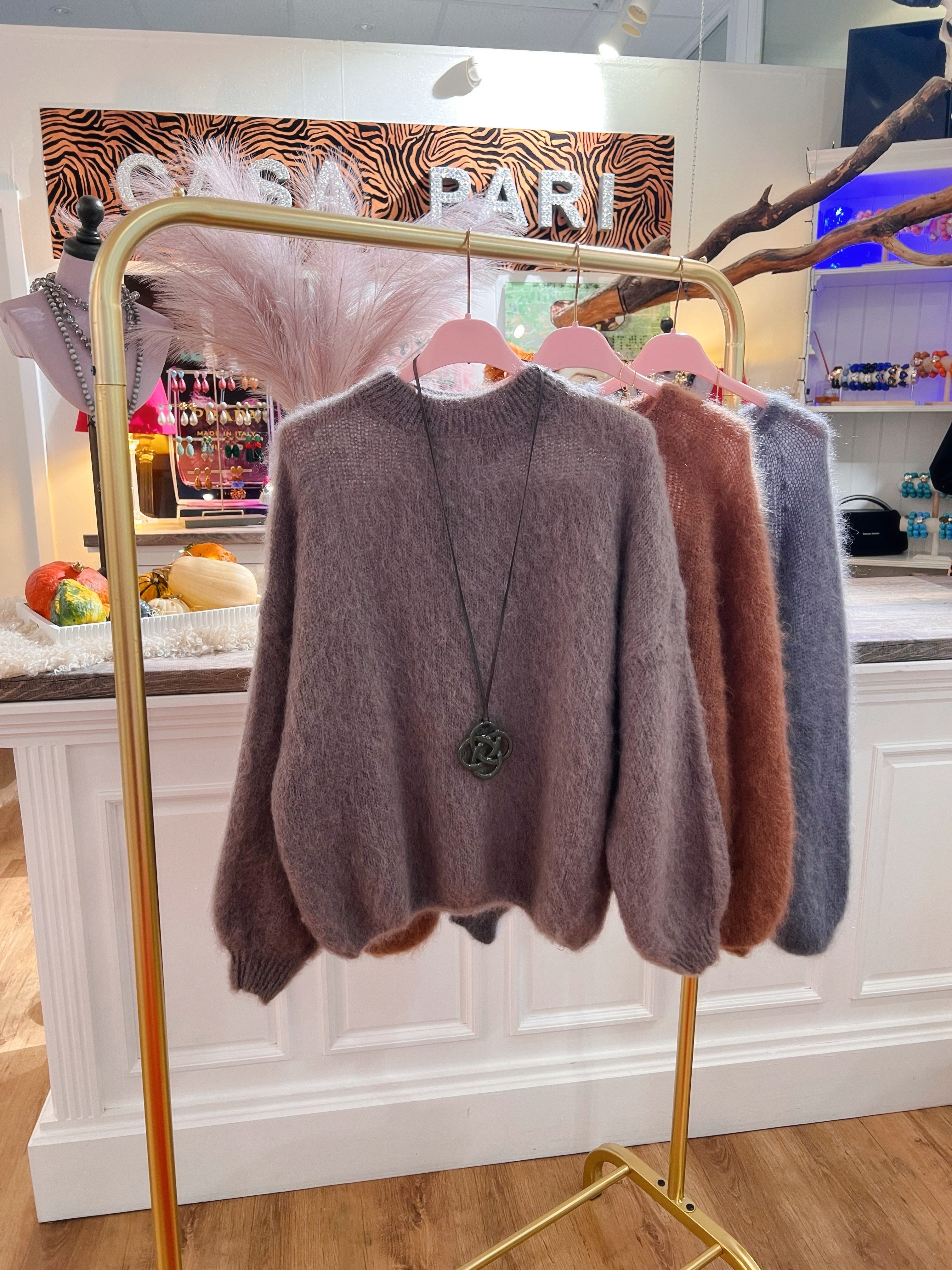 Drei weiche Pullover aus Kid Mohair in warmen Herbsts Tönen. Taupe mit Kette, Karamell und Grau. Hängen auf roséfarbenen Kleiderbügel an einer goldenen Stange in einer Boutique.