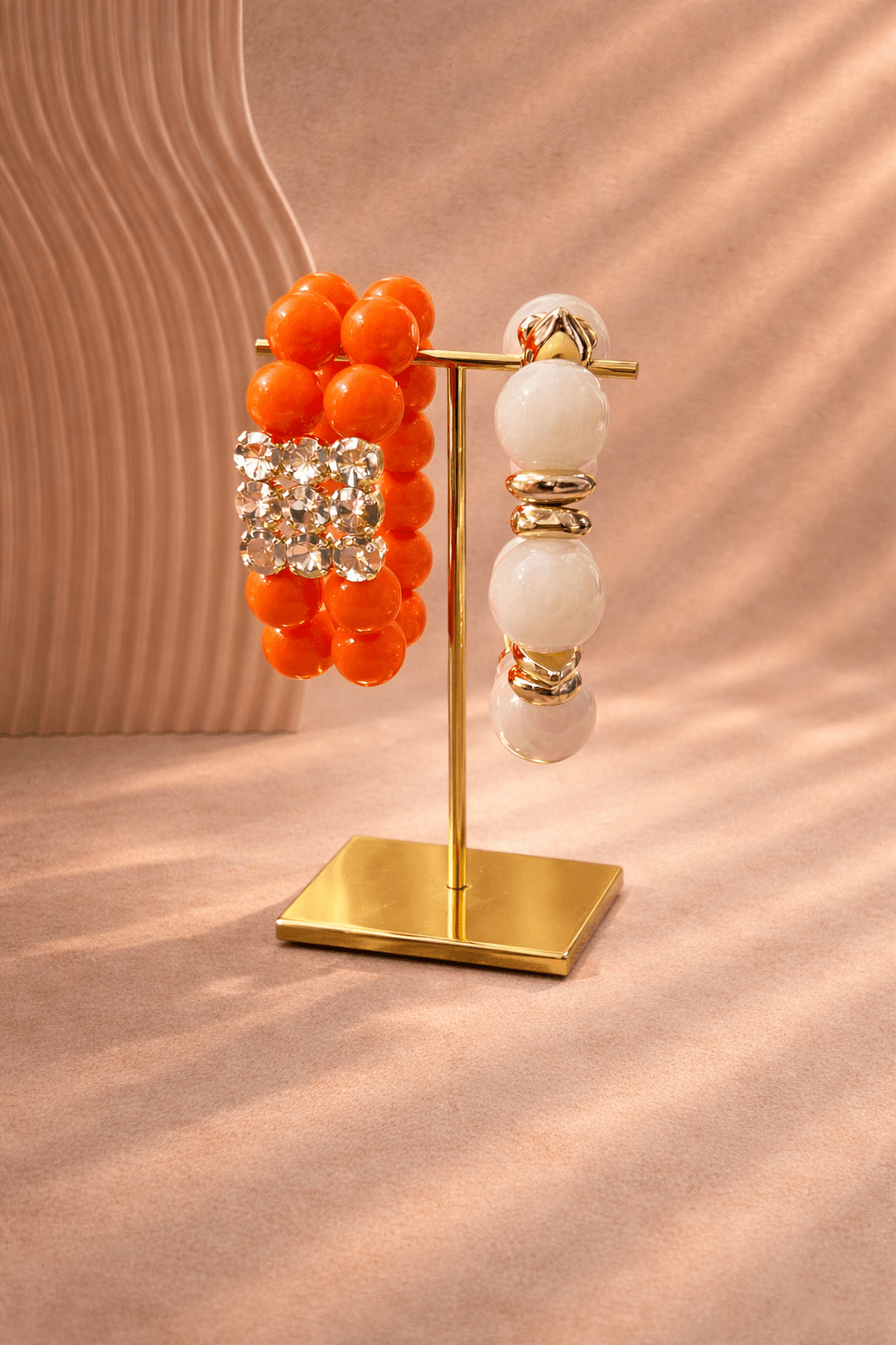 Armband-Set auf goldenem Schmuckständer vor beigem Hintergrund: links mehrere orangefarbene Perlenarmbänder mit quadratischem Strass-Element, rechts ein weißes Perlenarmband mit goldfarbenen Zwischenelementen.