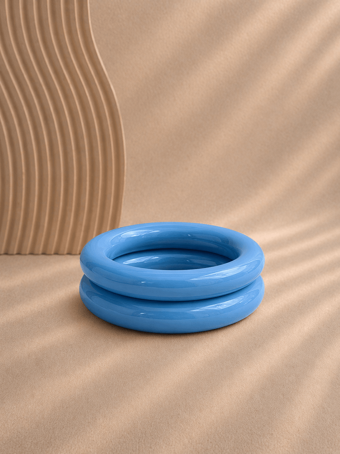 Blauee, glänzender Resin-Armreif liegt mittig auf warmem, sandfarbenem Hintergrund; links im Bild ein geriffeltes Deko-Element, weiches Licht und klare Darstellung.