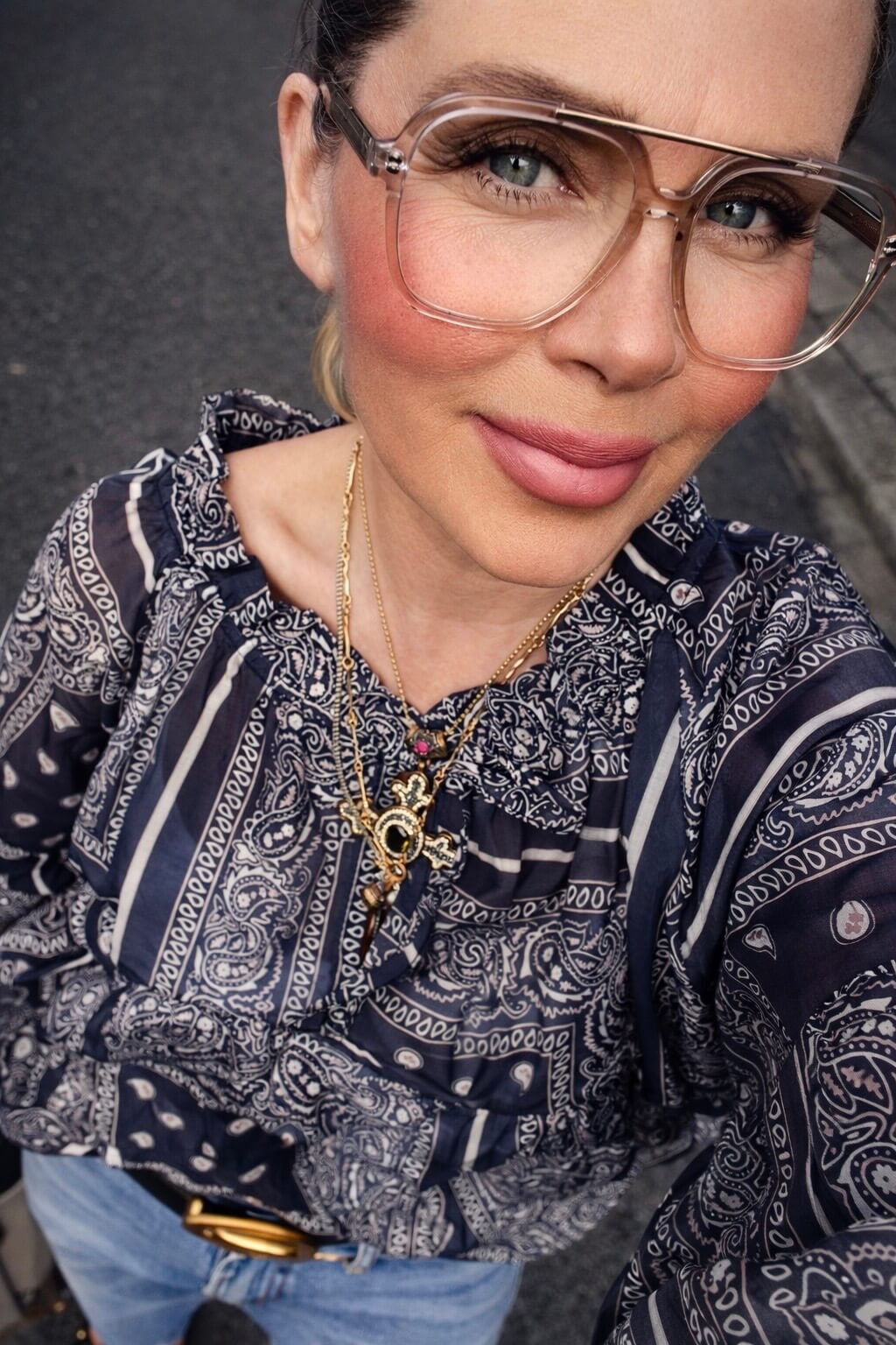 Frau trägt blaue Paisley-Bluse mit Jeans und goldener Halskette im Sonnenlicht auf einer Straße.