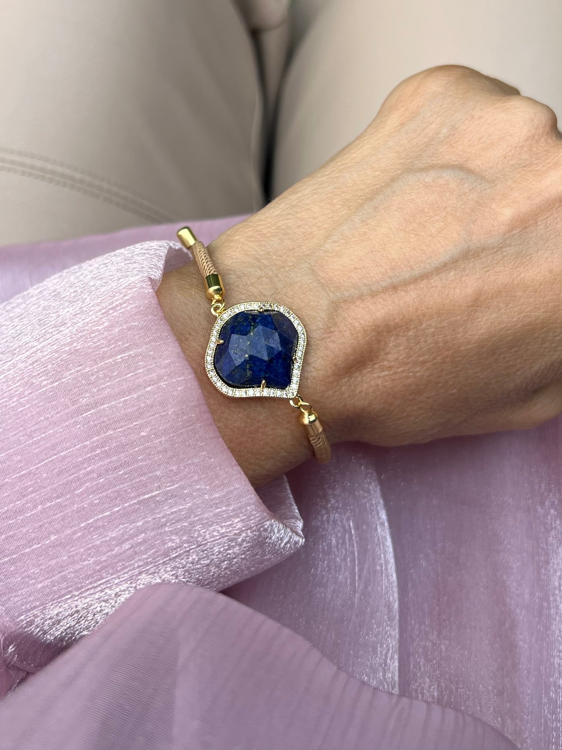 Beiges Armband mit blauem Schmuckstein an Hand getragen. Auf rosa Stoff fotografiert.