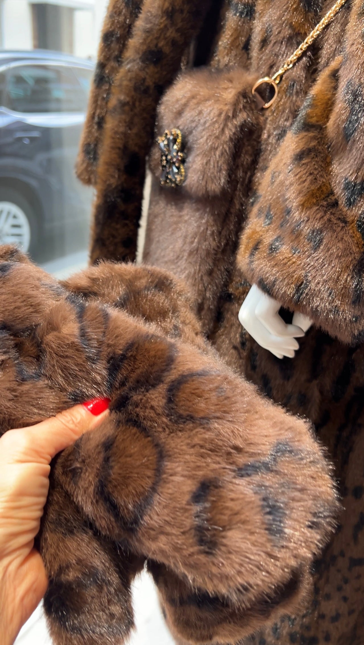 Nahaufnahme eines braunen Kunstpelzmantels mit Leopardenmuster. Eine Hand hält den passenden Handschuh davor. Im Hintergrund eine Schaufensterpuppe mit derselben Jacke und kleiner brauner Tasche mit goldener Brosche zu shene.