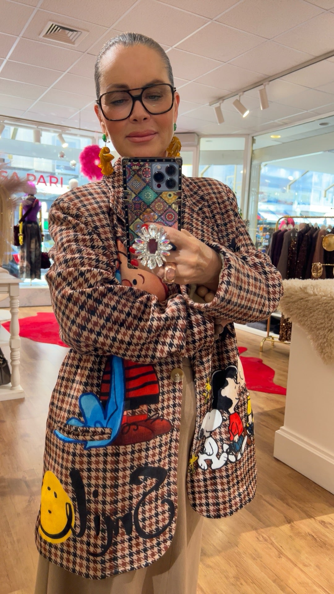 Nahaufnahme derselben Person im Boutique-Store. Der karierte Blazer mit bunten Comic-Motiven ist im Fokus. Die Person hält ein Smartphone und trägt große Brille, auffällige Ohrringe und einen großen, glitzernden Ring. Im Hintergrund sind Kleiderständer und Dekoelemente zu sehen.