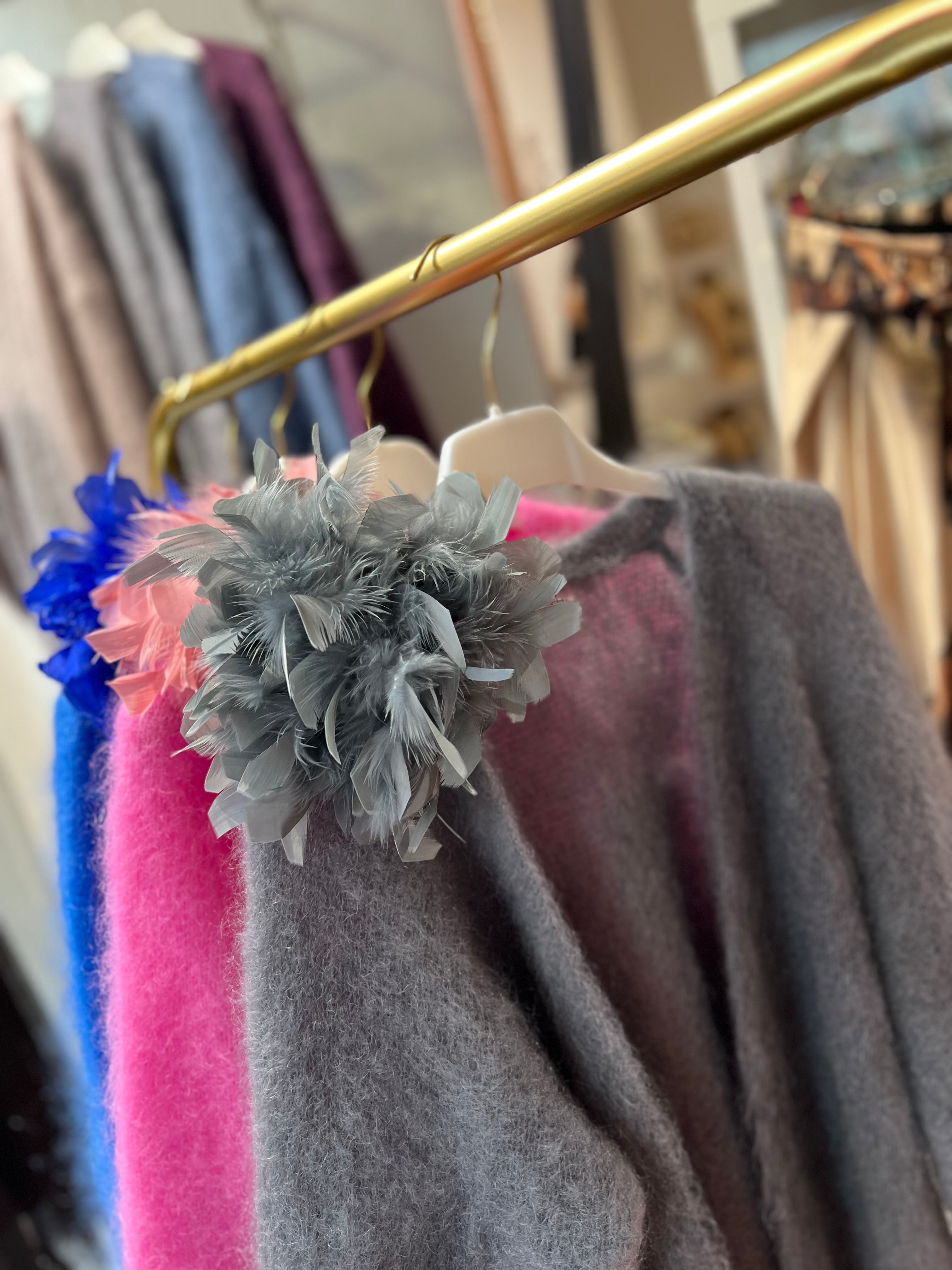 Drei Federbroschen mit großen Blüten aus Federn, eine in Grau und eine in Rosa und leuchtendem Blau.. befestigt an einem grau, pink und blauem Pullover auf einem goldenen Kleiderständer.