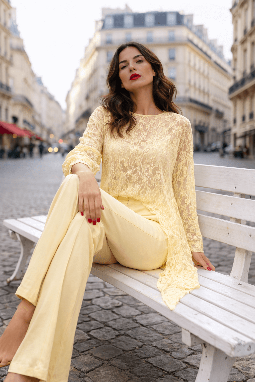 Frau sitzt auf einer weißen Holzbank in einer Pariser Innenstadtstraße mit Kopfsteinpflaster und hellen Altbauten im Hintergrund. Sie trägt ein vanillefarbenes, asymmetrisches Spitzenoberteil mit langen Ärmeln sowie eine weite, hellgelbe Hose.