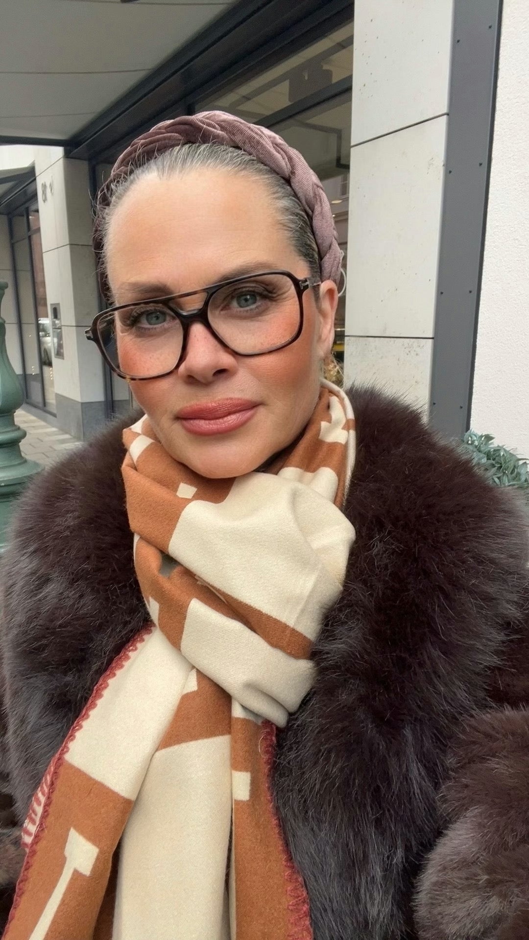 Frau trägt hellbraunen Haarreifen dazu große Brille. Dazu einen Camel beigefarbenen Schal. Und ein Fake Fur Pelzjacke.