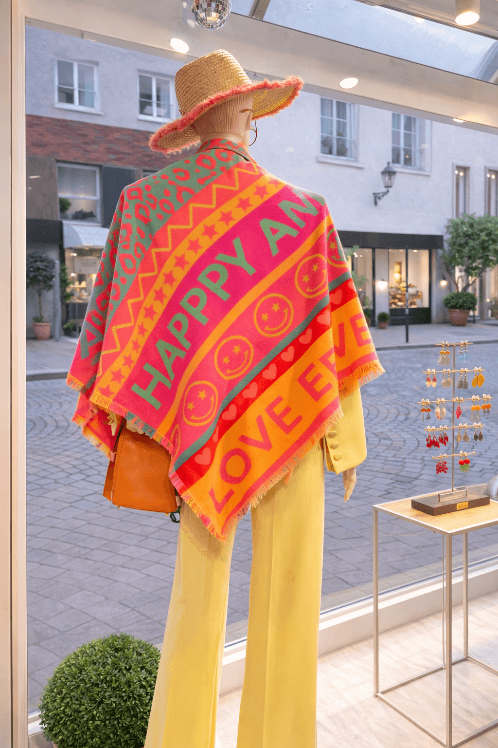 Schaufensterpuppe trägt ein buntes Tuch mit den Schriftzügen „Happy“ und „Love“ sowie Smiley- und Herzmotiven in Pink, Orange und Grün. Kombiniert mit gelber Hose und Strohhut in einem hellen Innenraum vor einer Fensterfront.