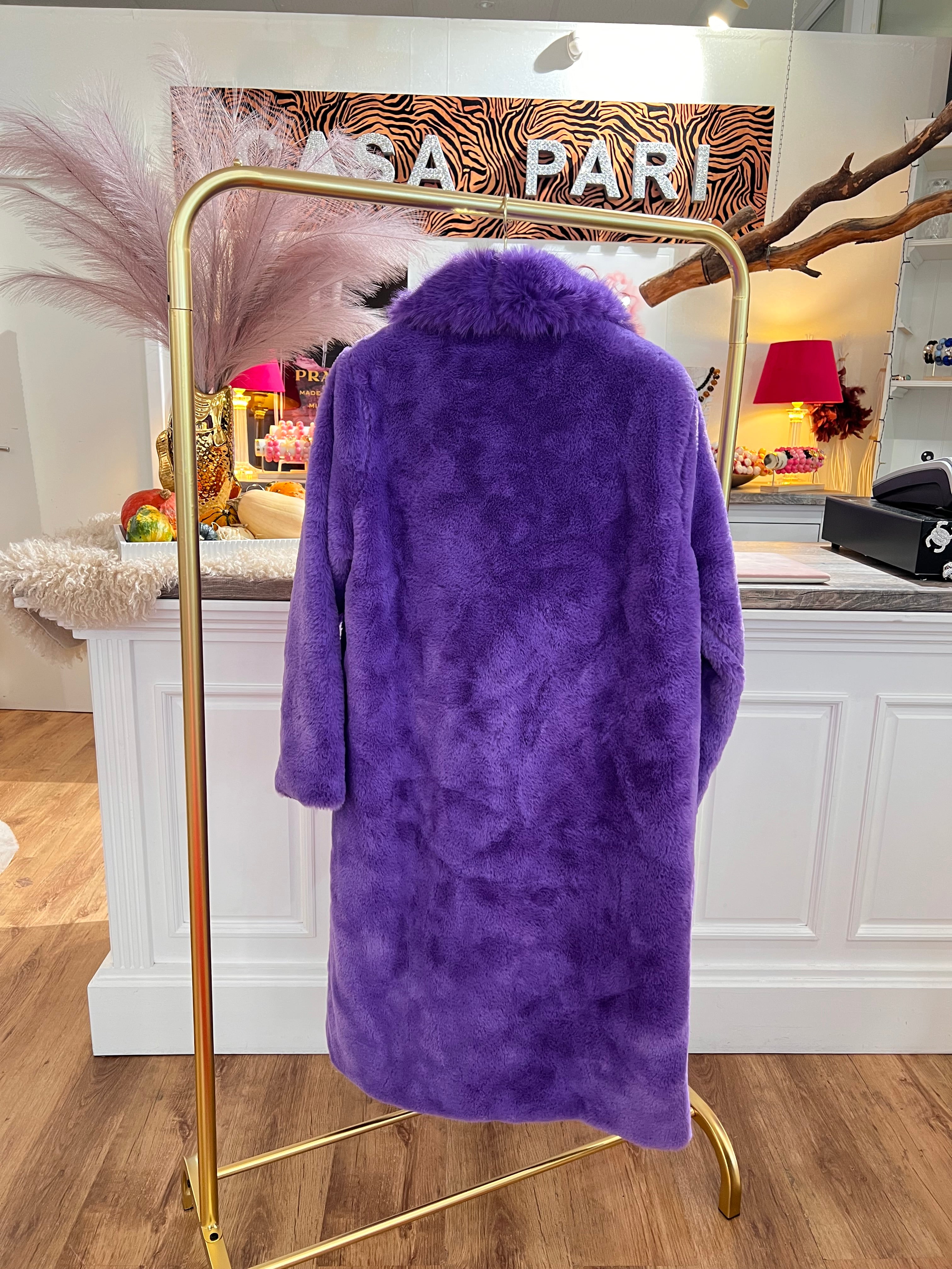 Rückansicht eines langen flauschigen Mantels in tiefen Violett hängt auf einem goldenen Kleiderständer vor einer weißen Theke.