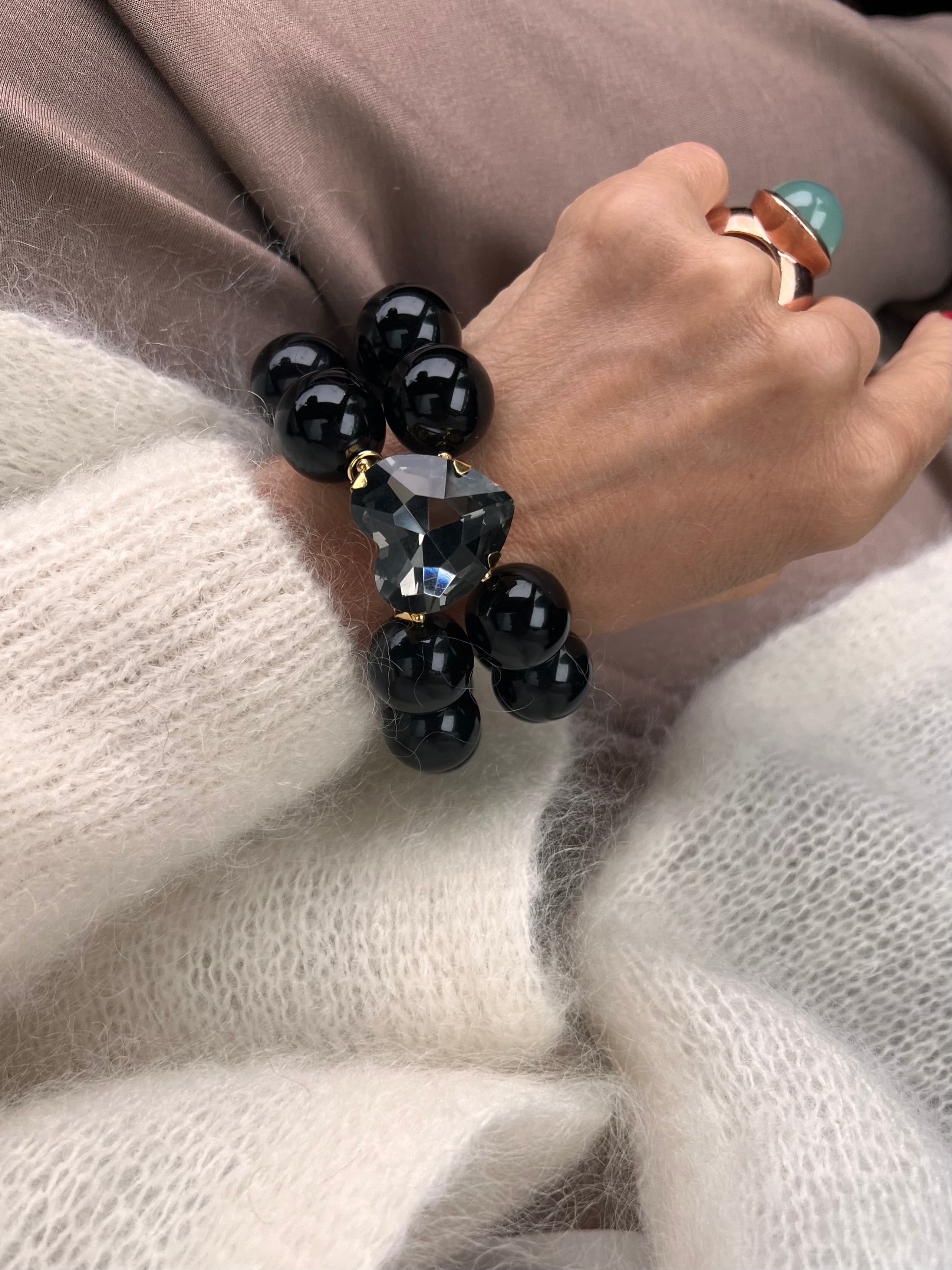 Armband in Schwarz mit großem Herzschmuckstein in Grau, und großem Ring. Am Handgelenk auf beigem Stoff fotografiert.