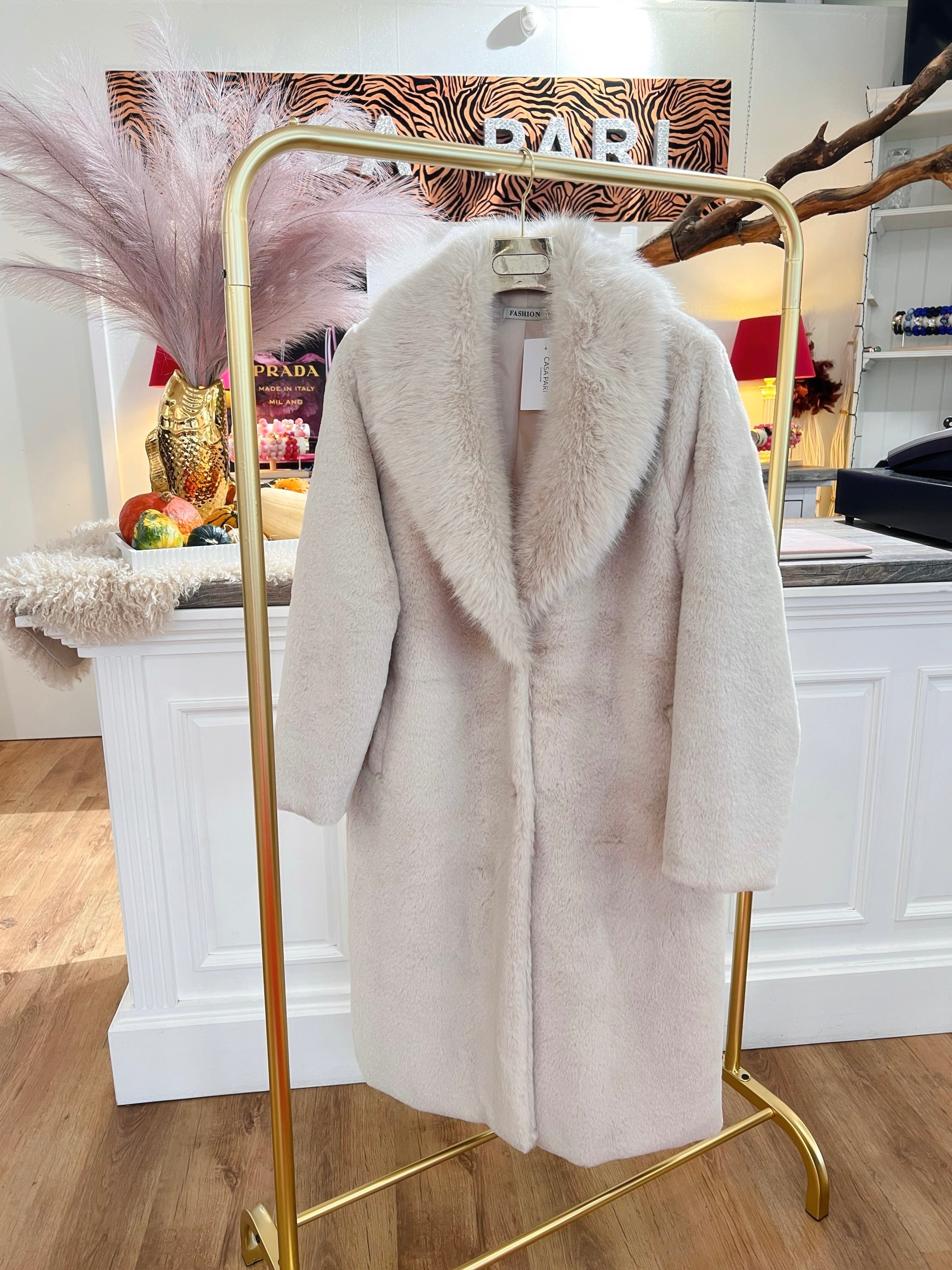 Langer, flauschiger Mantel in Creme hängt auf einem goldenen Kleiderständer vor einer weißen Theke.