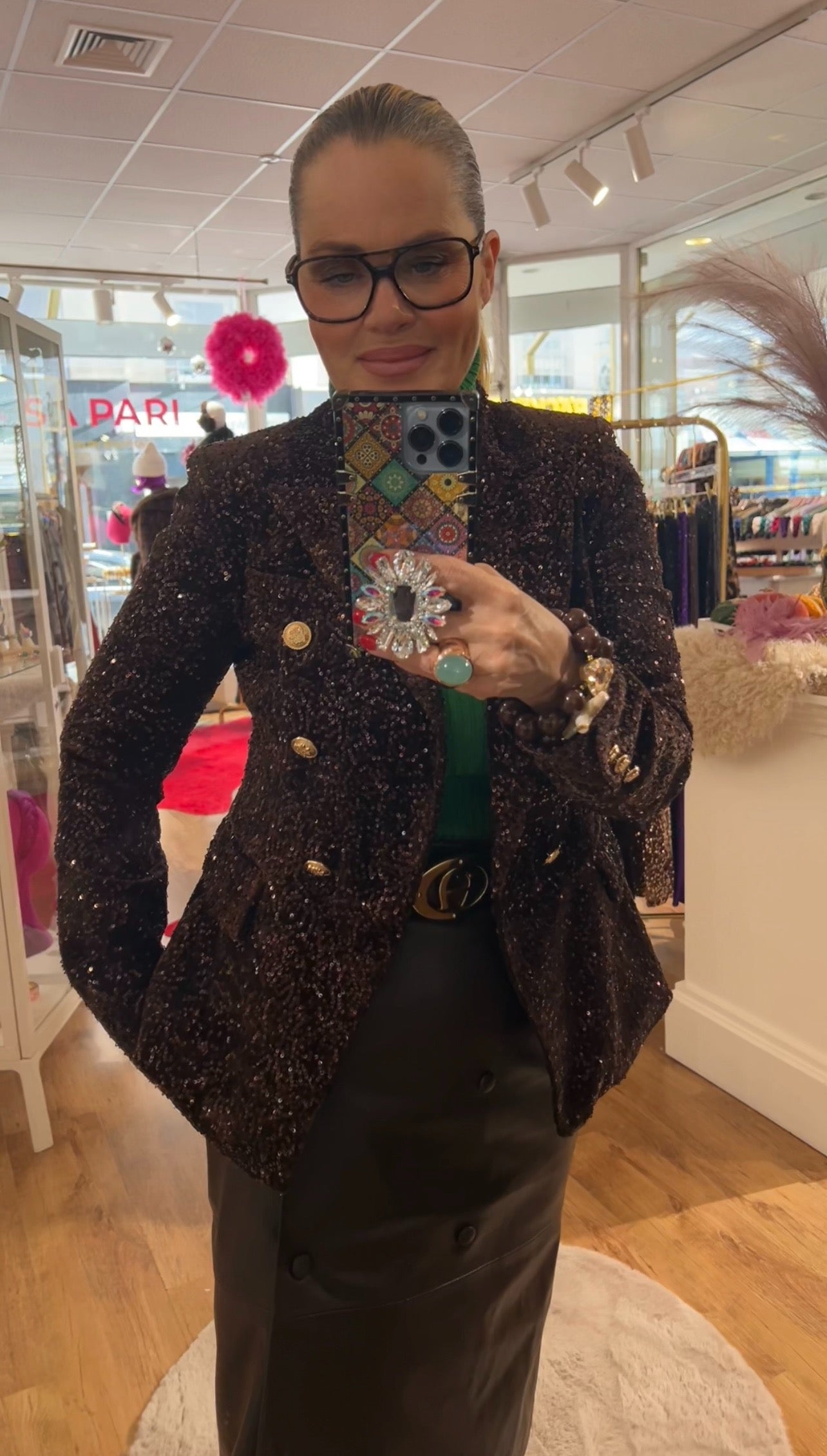 Frau steht im Laden vor einem Spiegel und macht ein Spiegel Selfie. Sie trägt einen dunklen schimmernden Pailletten Blazer mit goldfarbenen Knöpfen, einen grünen Rollkragenpulli und einen schwarzen Rock. In der Hand hält sie ein Smartphone mit gemustertem Case und großen Ring. Im Hintergrund eine Ladeneinrichtung.