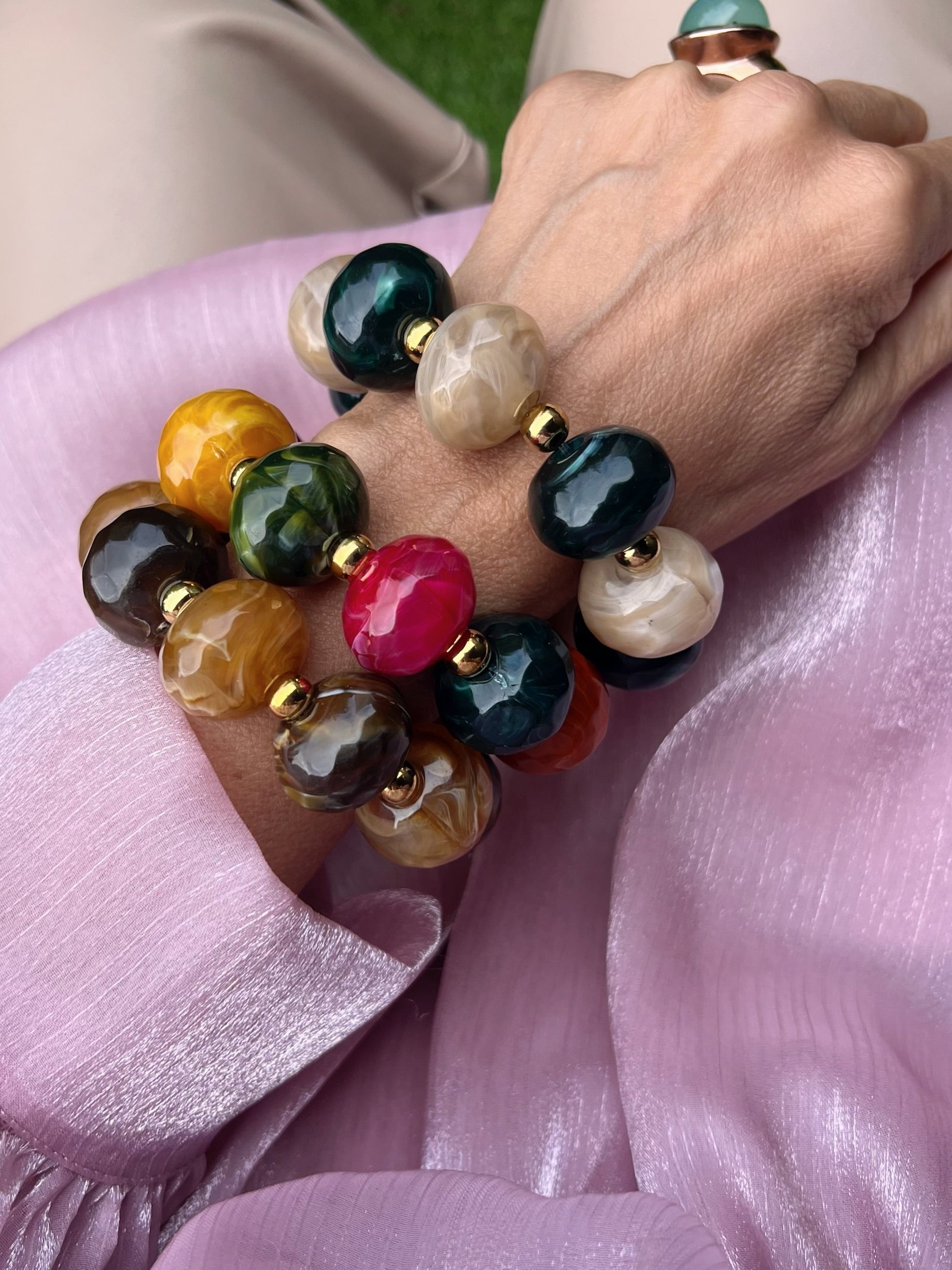 Drei große bunte Armbänder an einem Handgelenk fotografiert auf Rosa Stoff.