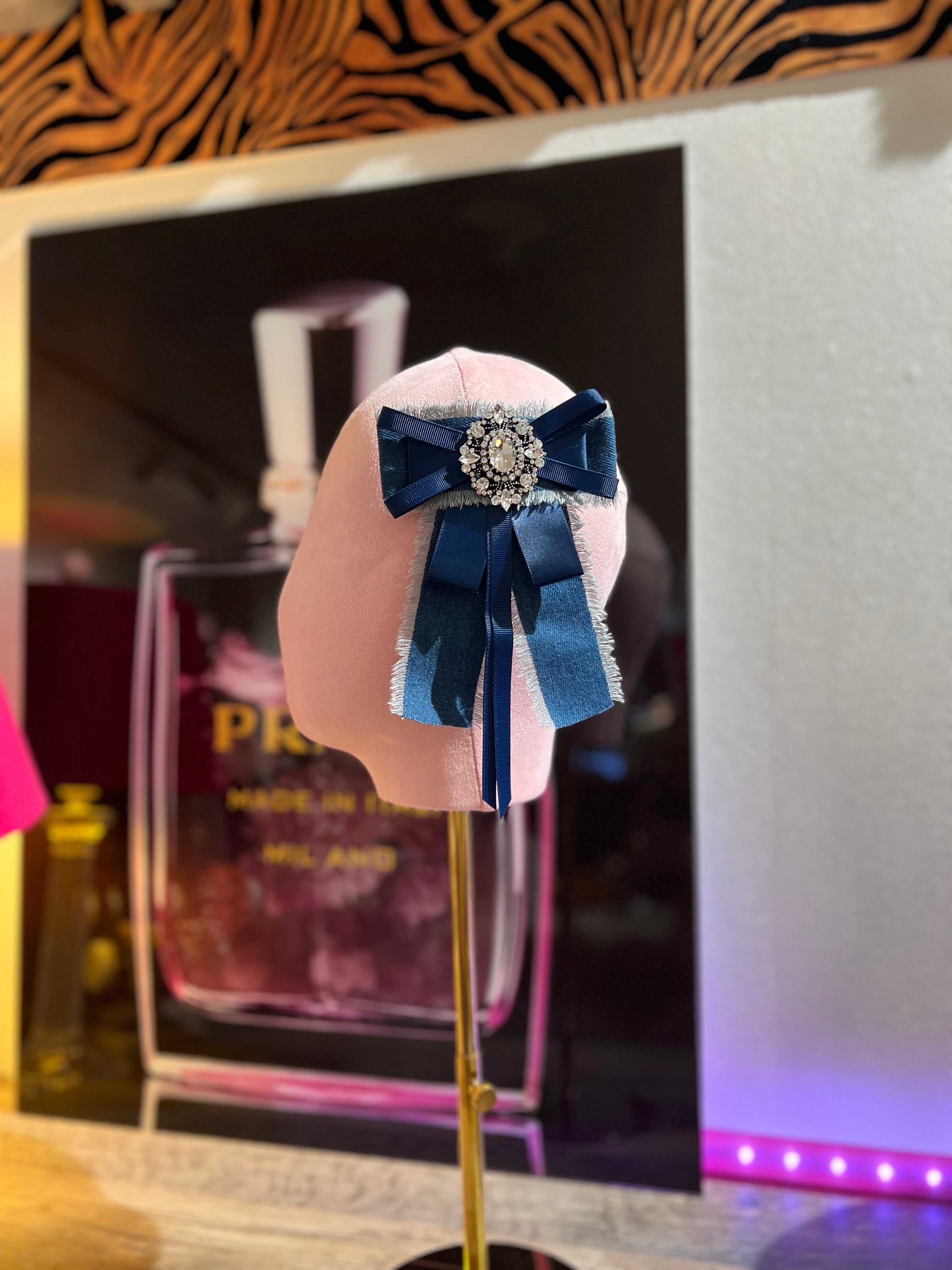 Jeansbrosche mit dunkelblauer Schleife und Strassverzierung, befestigt auf einer rosa Stoffpuppe vor einem dekorativen Hintergrund mit Parfüm Flakon Motiv.