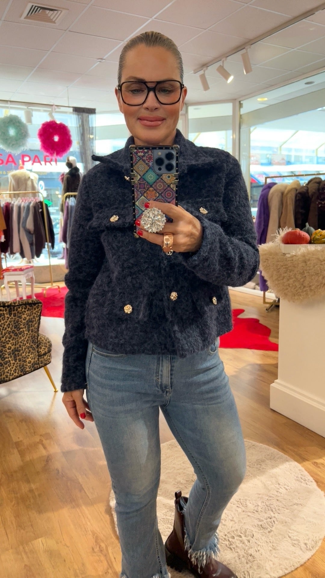 Frau trägt blaue Jacke mit goldenen Knöpfen dazu eine Jeans und braune Boots. Sie hält ein Handy in der Hand, trägt einen großen Ring, fotografiert in einer Boutique.