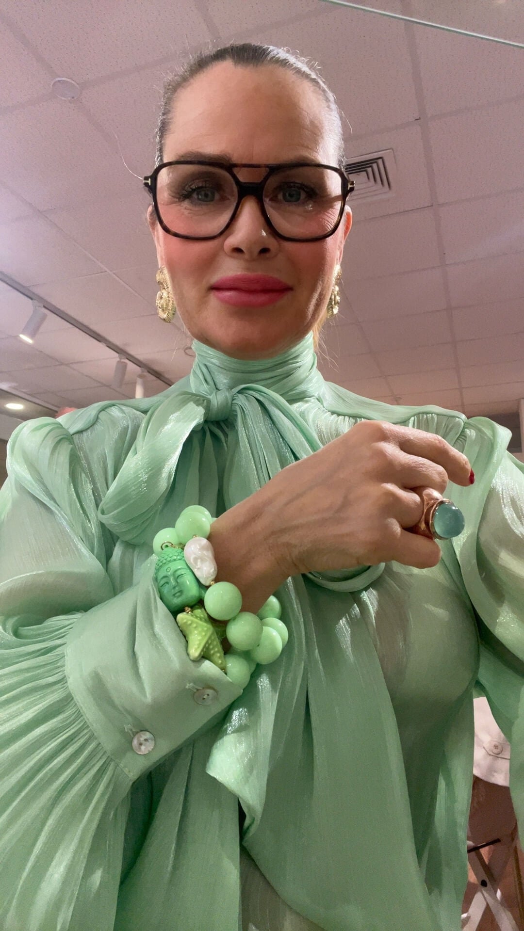 Person trägt eine mintgrüne, transparente Bluse mit Schleifenkragen, dazu ein Armband aus mintgrünen Perlen mit grünem Buddha-Element, weißer Perle und kleinem Seestern-Anhänger am Handgelenk. Ergänzt durch eine grüne Ringfassung mit rundem Stein und goldfarbene Ohrringe, aufgenommen in einem Innenraum.
