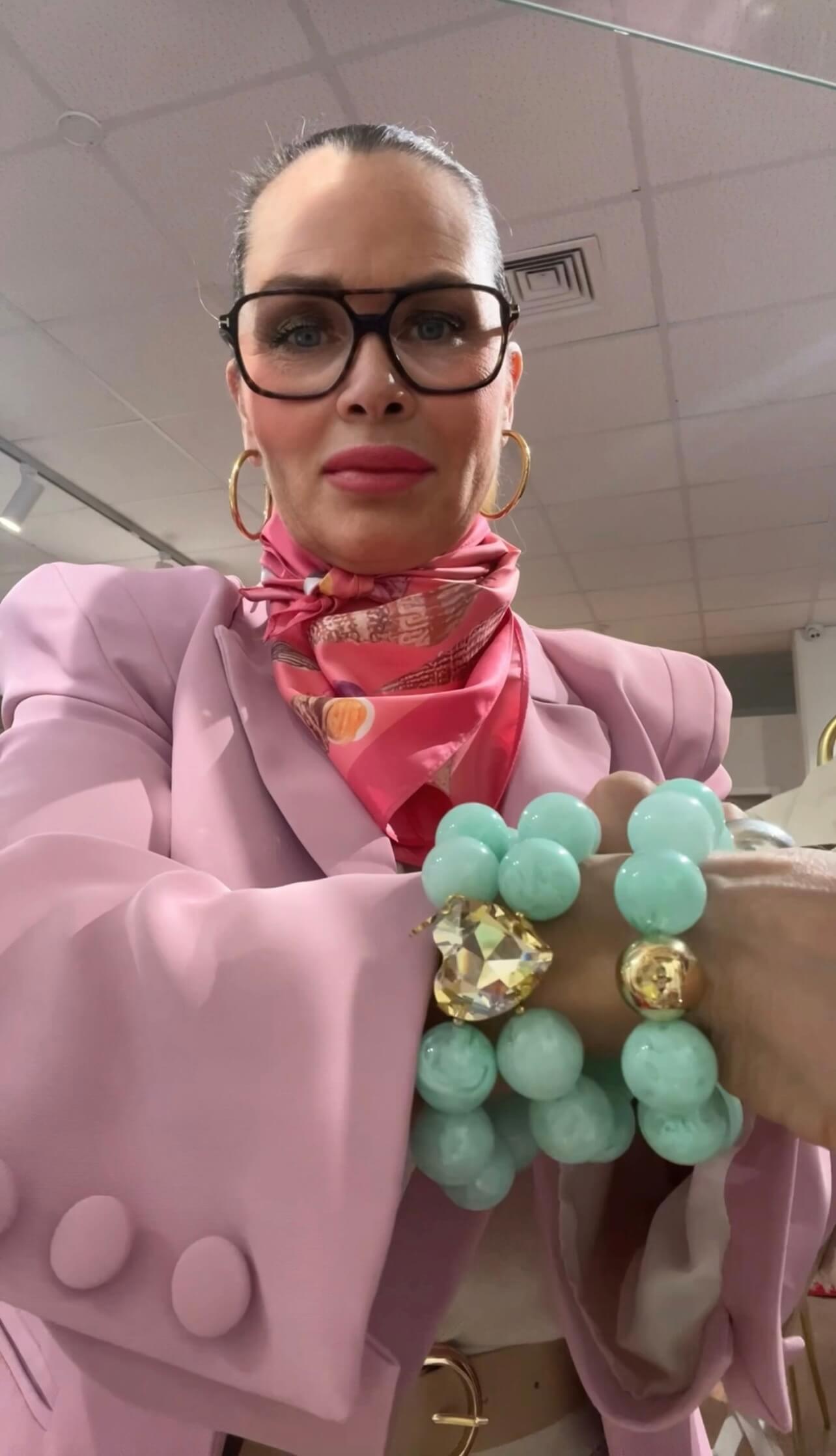 Person mit hochgebundenem Haar trägt eine rosafarbene Jacke, eine rosa gemusterte Halstuchkrawatte, eine dunkle Brille und goldene Creolen. Am Handgelenk sind zwei Armbänder mit großen mintfarbenen Perlen, einem goldfarbenen Element und einem facettierten Schmuckstein zu sehen.