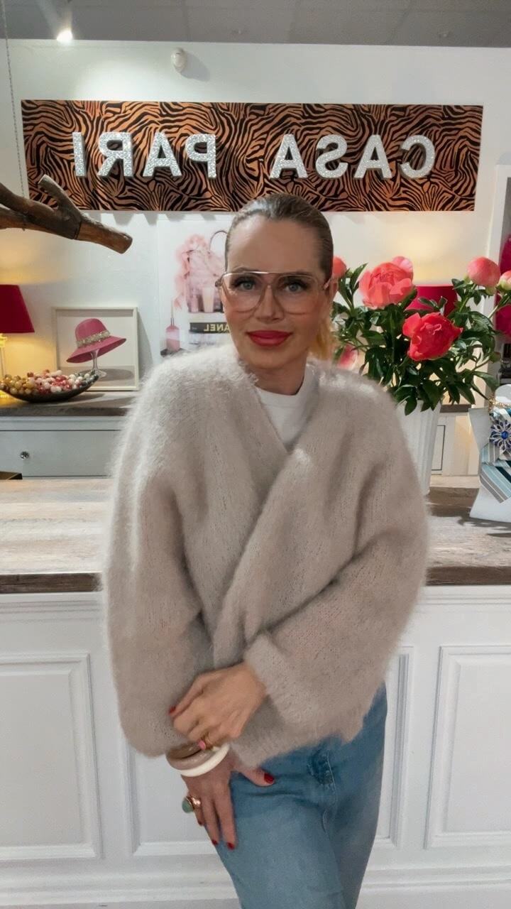 Frau trägt einen hellen, weichen Strick-Cardigan in Wickeloptik mit Jeans und Schmuck, frontal stehend in einer Boutique.