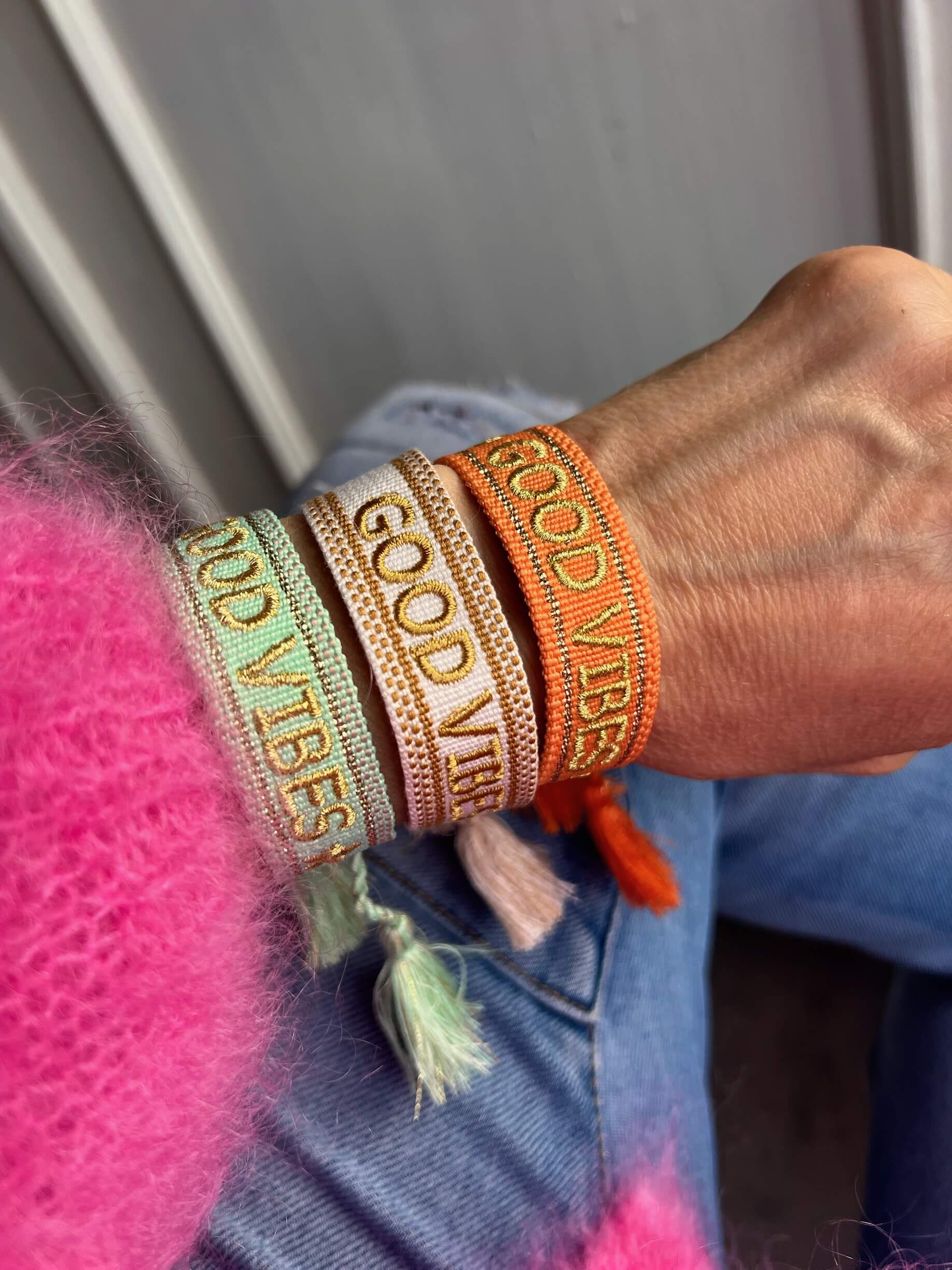 Gewebte Stoffarmbänder mit der Aufschrift „GOOD VIBES“ in Goldschrift, getragen am Handgelenk.
Farben: Mintgrün, Creme und Orange.
Jedes Armband mit seitlichen Quasten.
Person trägt einen pinkfarbenen, flauschigen Pullover und eine Jeans.