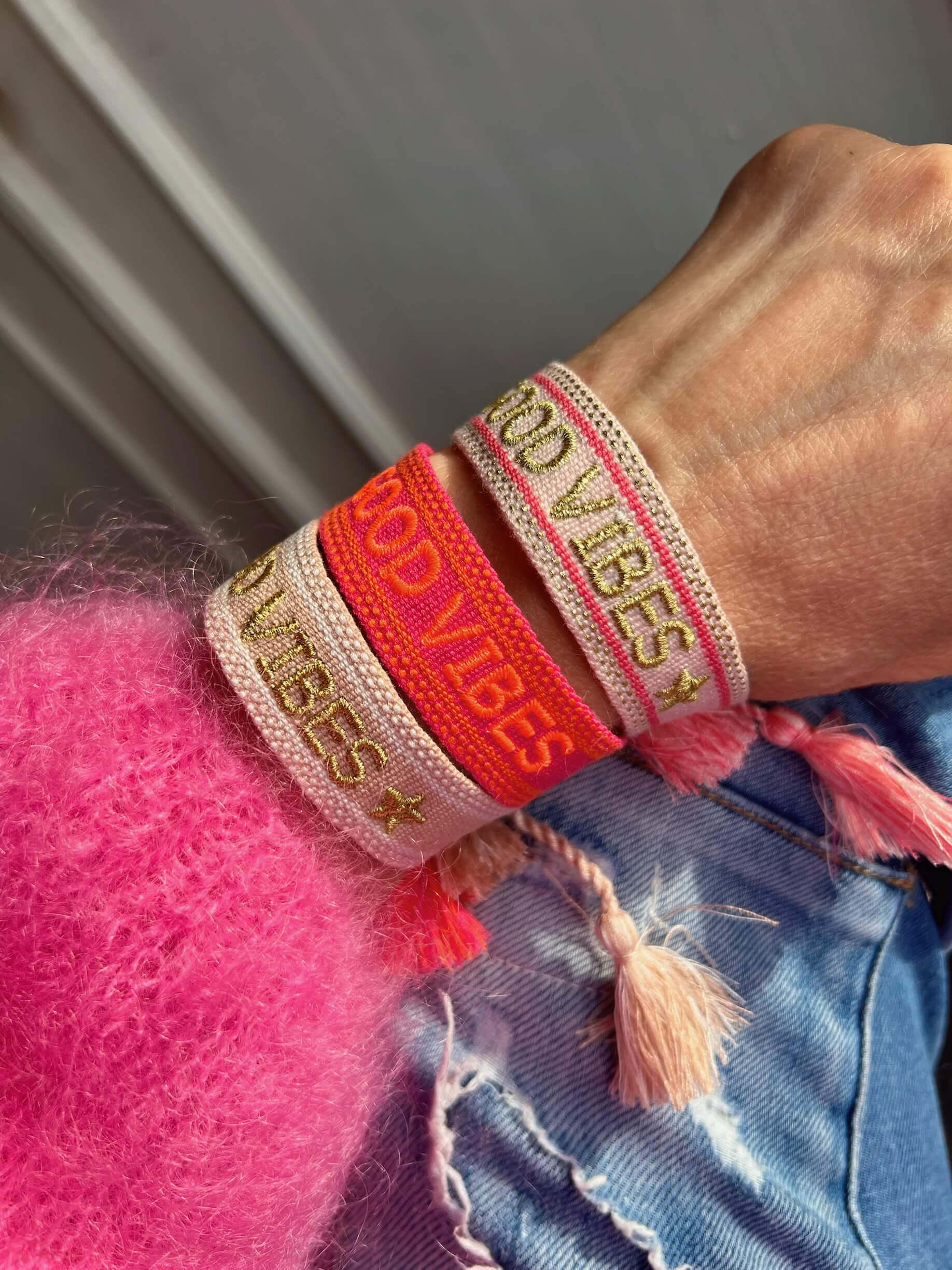 Handgelenk mit drei gewebten Stoffarmbändern in Pink-, Beige- und Koralltönen mit goldfarbener Schrift „GOOD VIBES“ und Quasten; getragen zu pinkem Pullover und heller Jeans.