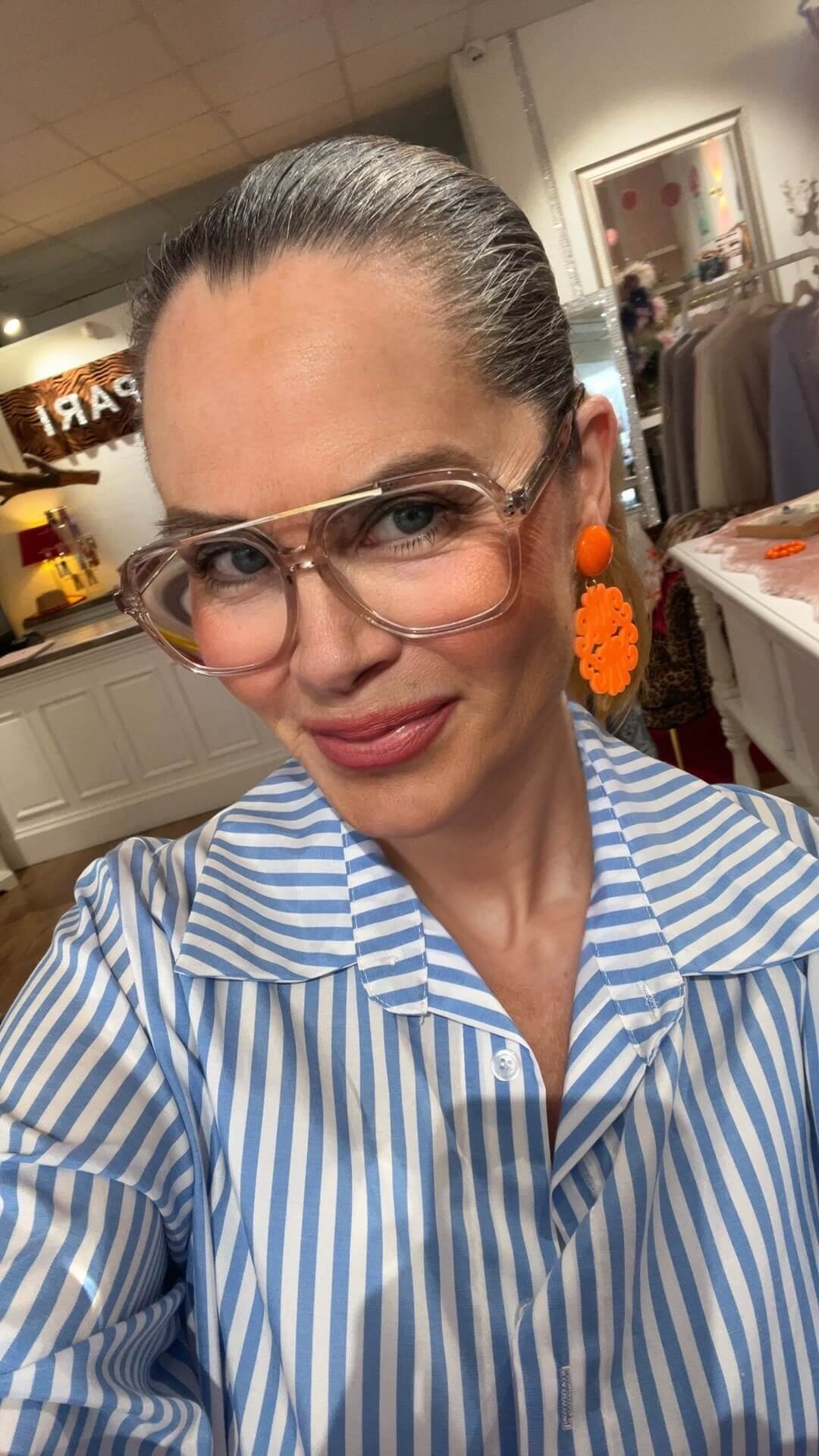 Frau mit zurückgebundenem Haar trägt eine blau-weiß gestreifte Bluse, transparente Brille und große orangefarbene Statement-Ohrringe, aufgenommen in einer Boutique mit Regalen und Spiegel im Hintergrund.