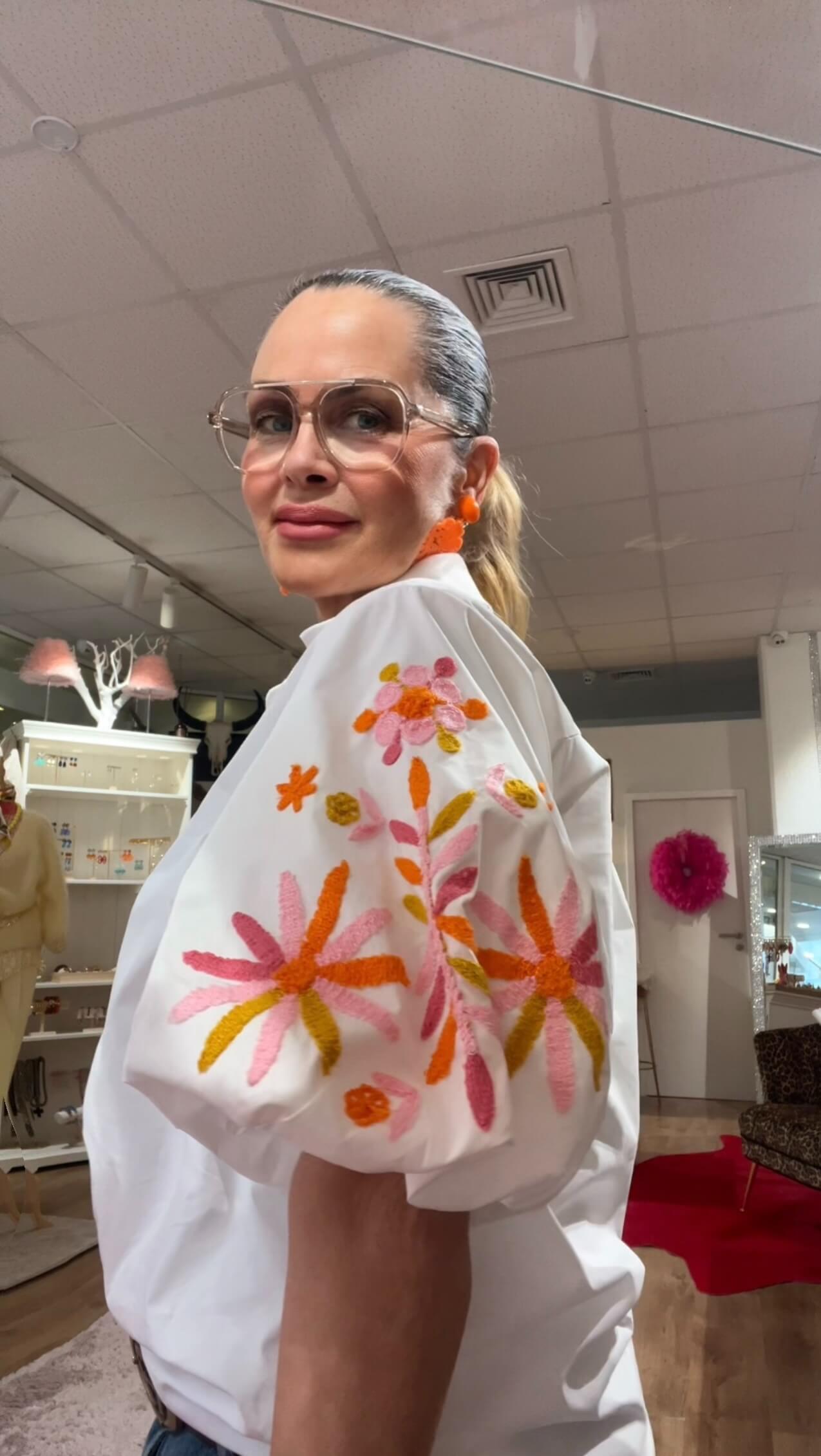 Frau mit zurückgebundenem Haar trägt eine weiße Bluse mit bunten Blumenstickereien am Ärmel, transparente Brille und orangefarbene Ohrringe, fotografiert in einer Boutique mit Regalen und Spiegel im Hintergrund.