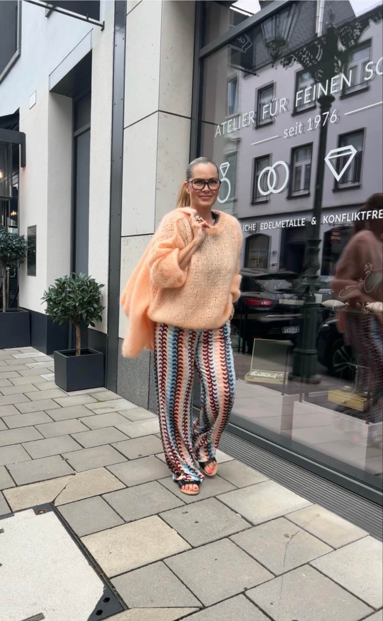 Person trägt gemusterte Palazzo-Hose in Türkis, Peach und Bordeaux mit weitem Bein und apricotfarbenem Strickpullover, aufgenommen auf einem Gehweg vor einem Schaufenster.
