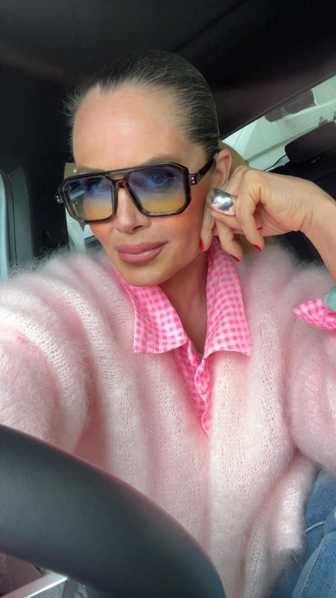 Frau mit dunklen Haaren trägt Oversize Sonnenbrille mit warm getönten Verlaufsgläsern und dunklem Schildpatt-Gestell. Sie sitzt im Auto und trägt eine rosa Mohair-Strickjacke über einer rosa-weiß karierten Bluse.