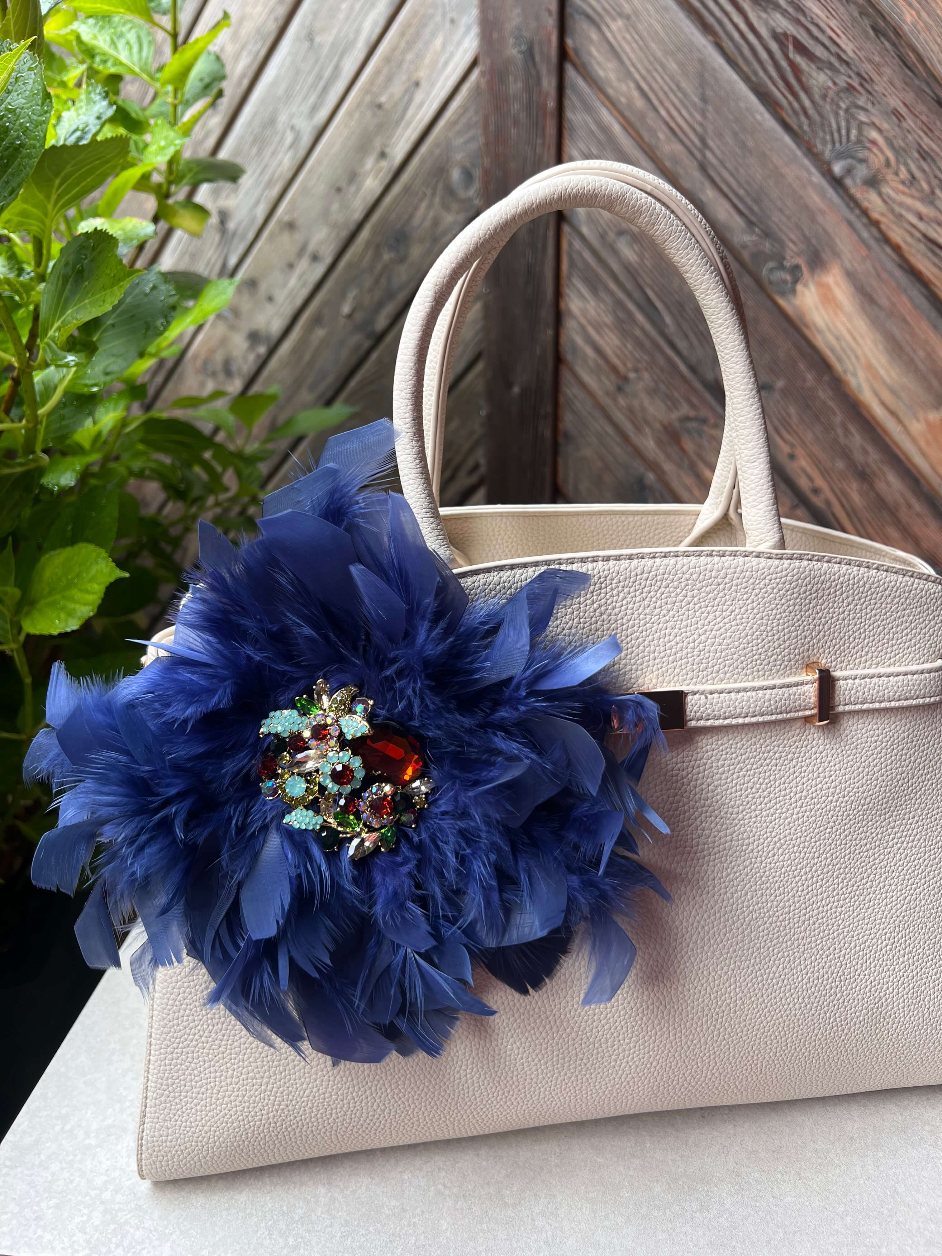 Blaue Brosche mit Schmuckelement an einer Beigen Tasche drapiert. Hinter einer Holzwand