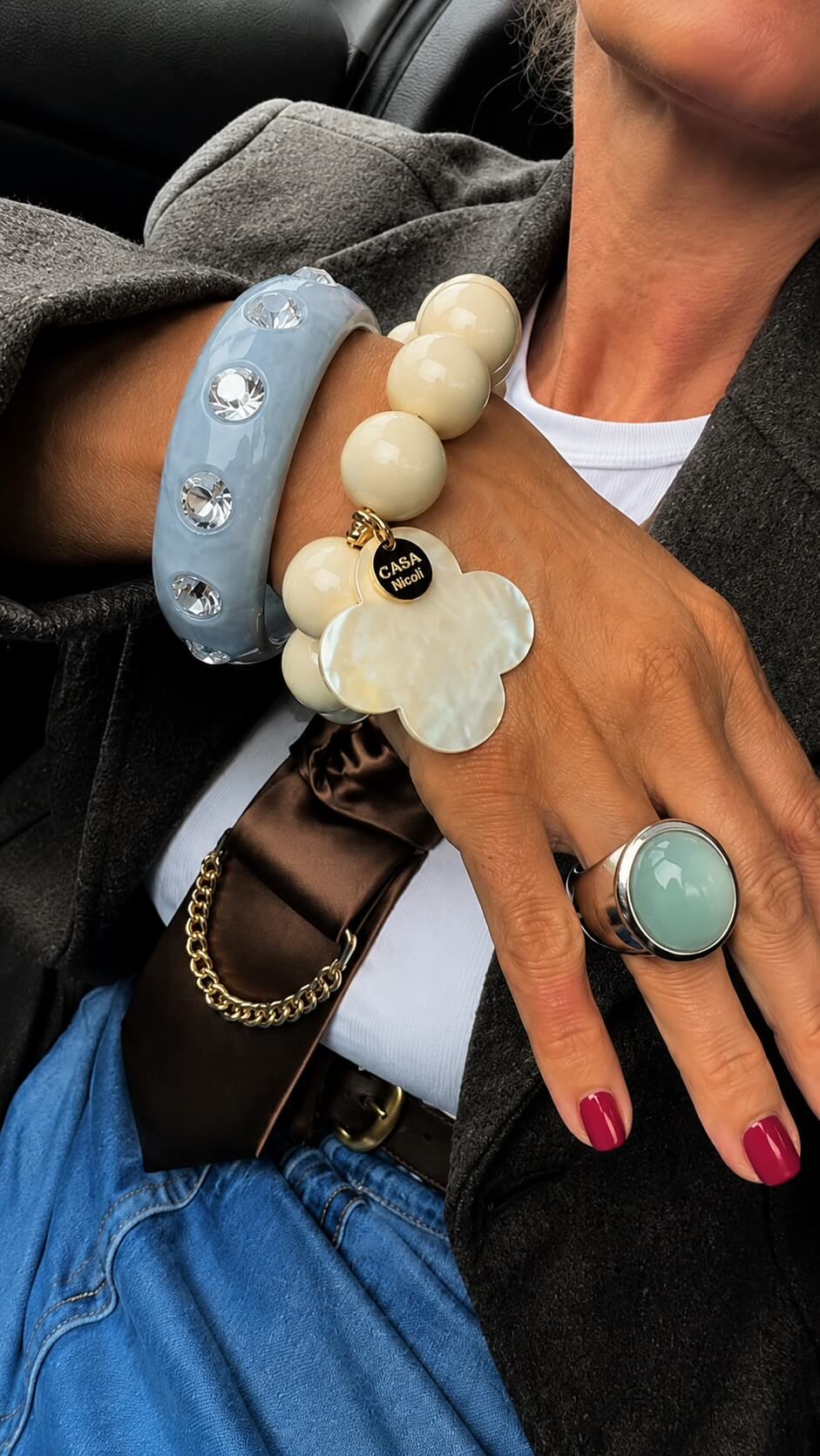 Handgelenk mit blauen Resina-Armreif mit Strass-Steinen, kombiniert mit einem cremefarbenen Perlenarmband und einem blauen Ring. Die Person trägt einen grauen Blazer, Jeans und eine braune Krawatte mit goldfarbener Kette.