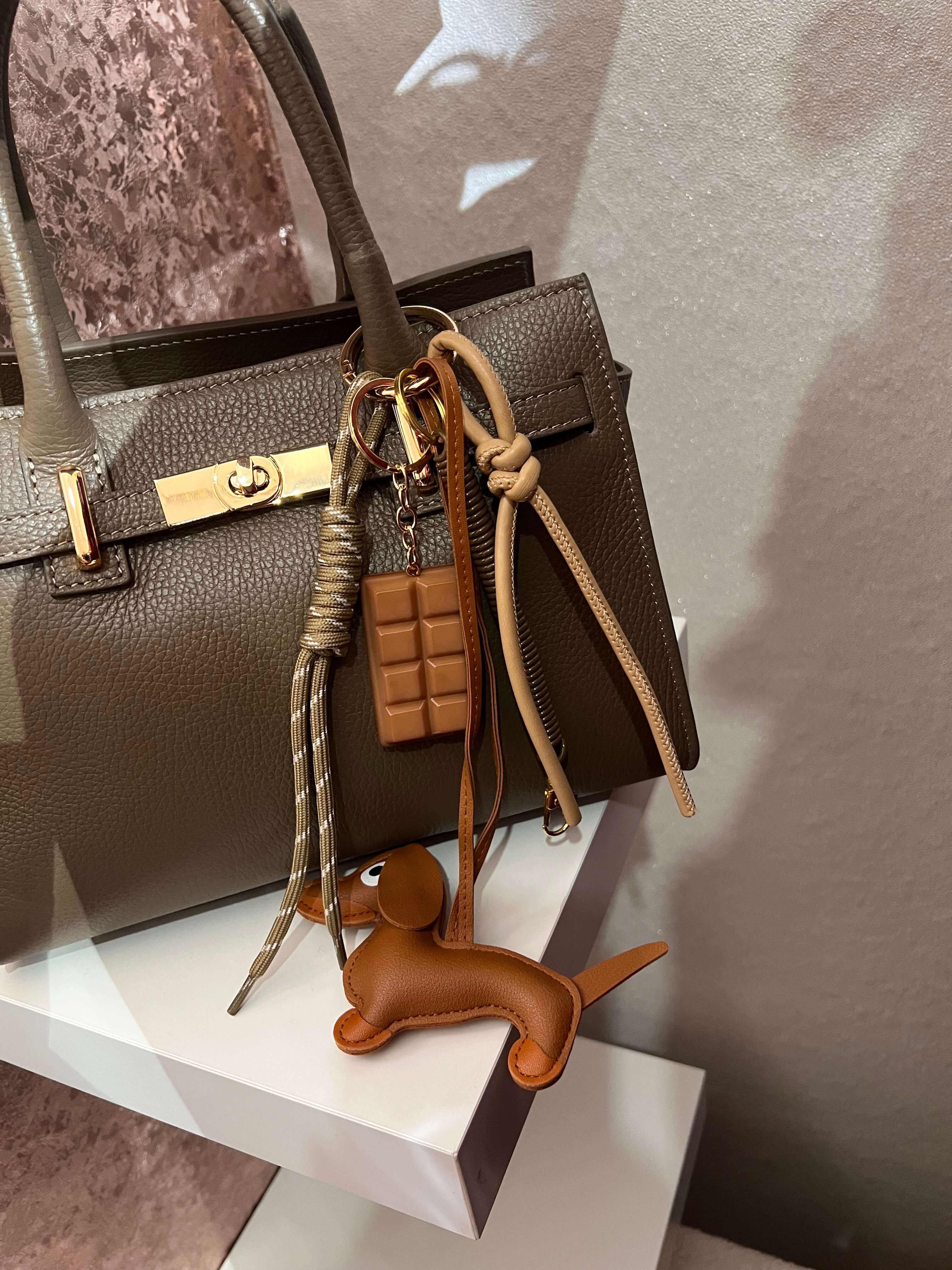Taschenanhänger in Form eines braunen Dackels aus Kunstleder, befestigt an einer camelfarbenen Handtasche mit goldfarbenem Clip und zusätzlichen dekorativen Anhängern.