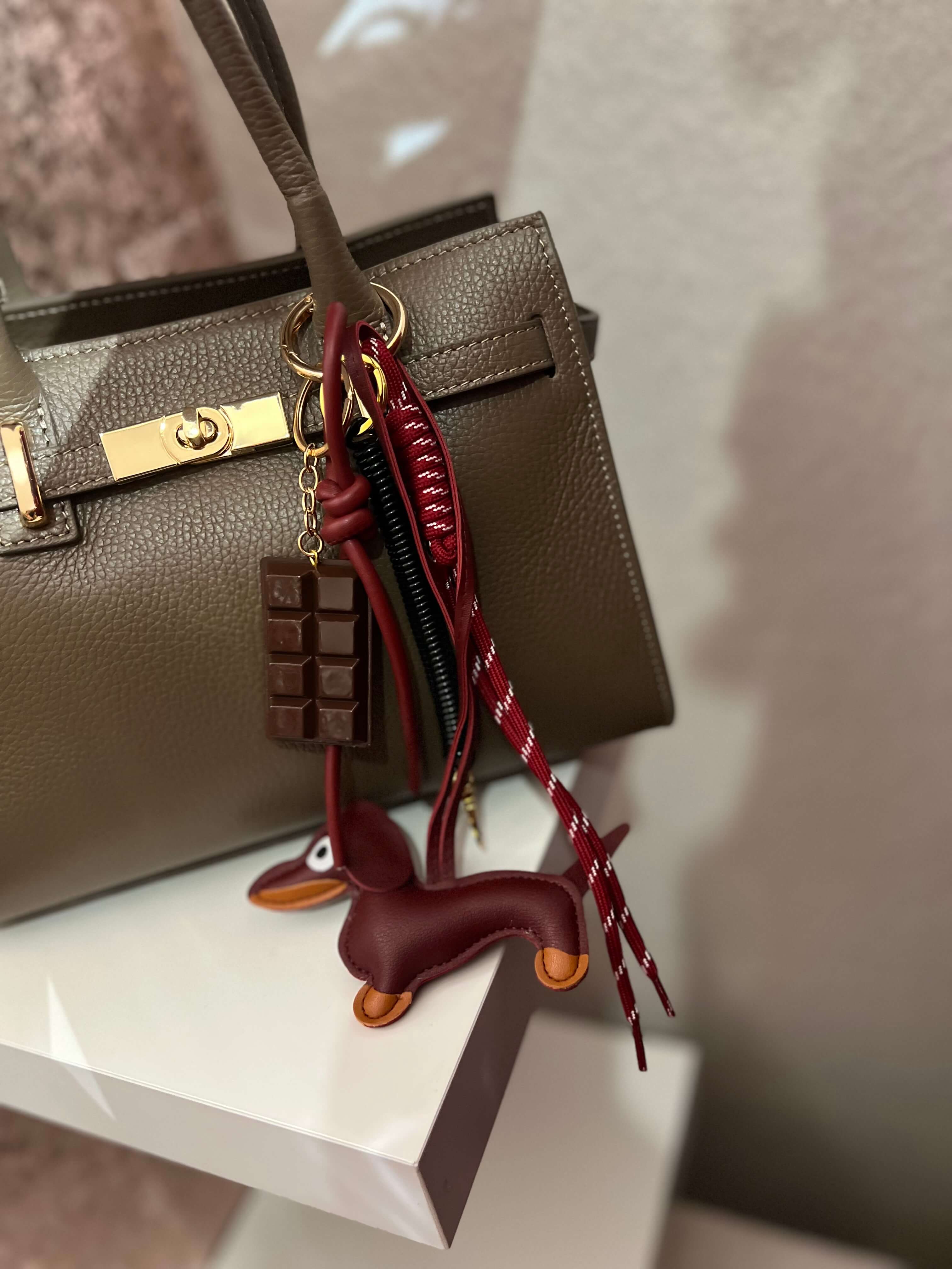 Taschenanhänger in Form eines braunen Dackels aus Kunstleder, befestigt an einer taupefarbenen Handtasche mit goldfarbenem Clip und zusätzlichen dekorativen Anhängern.