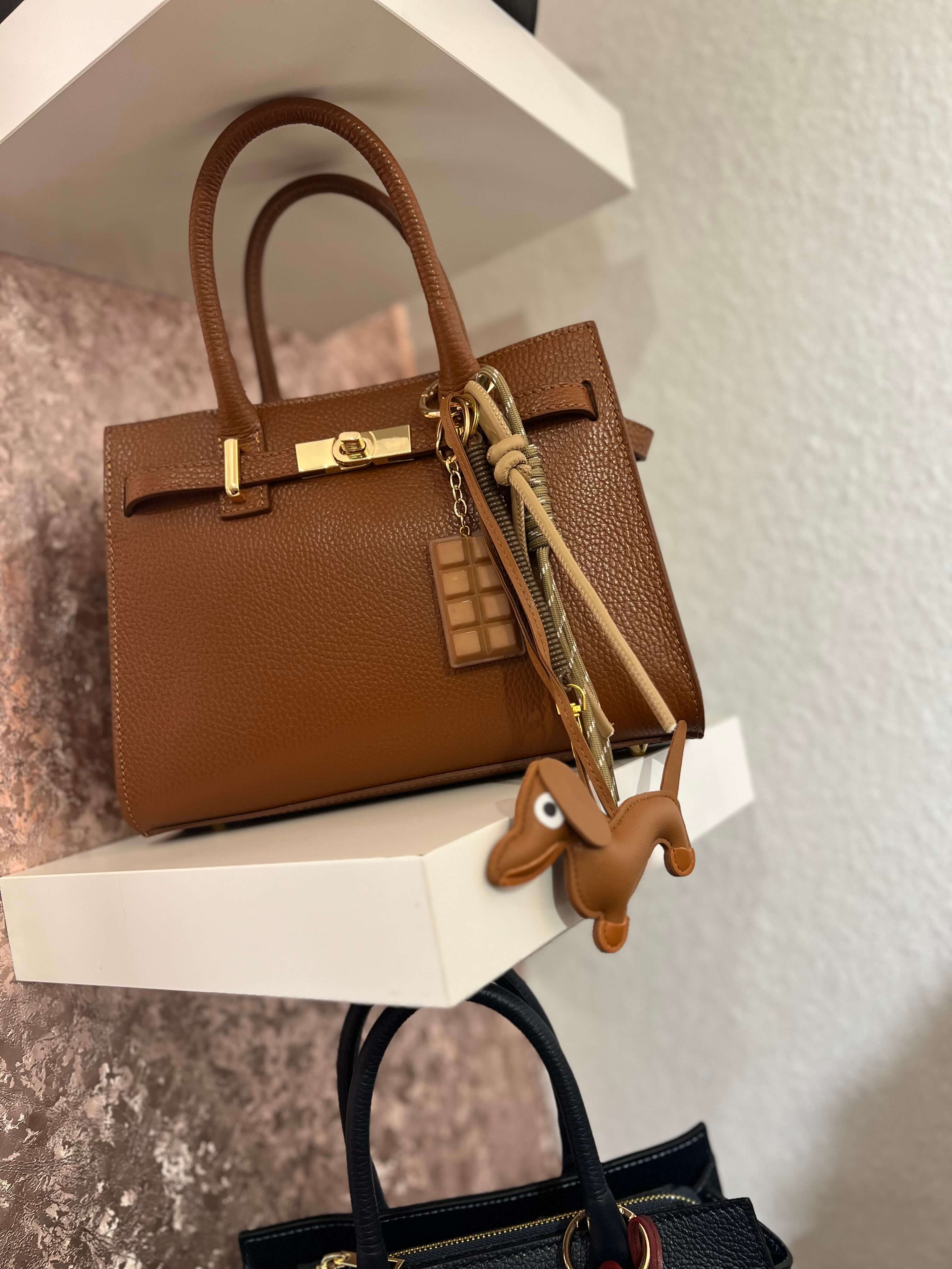 Taschenanhänger in Form eines braunen Dackels aus Kunstleder, befestigt an einer camelfarbenen Handtasche mit goldfarbenem Clip und zusätzlichen dekorativen Anhängern.