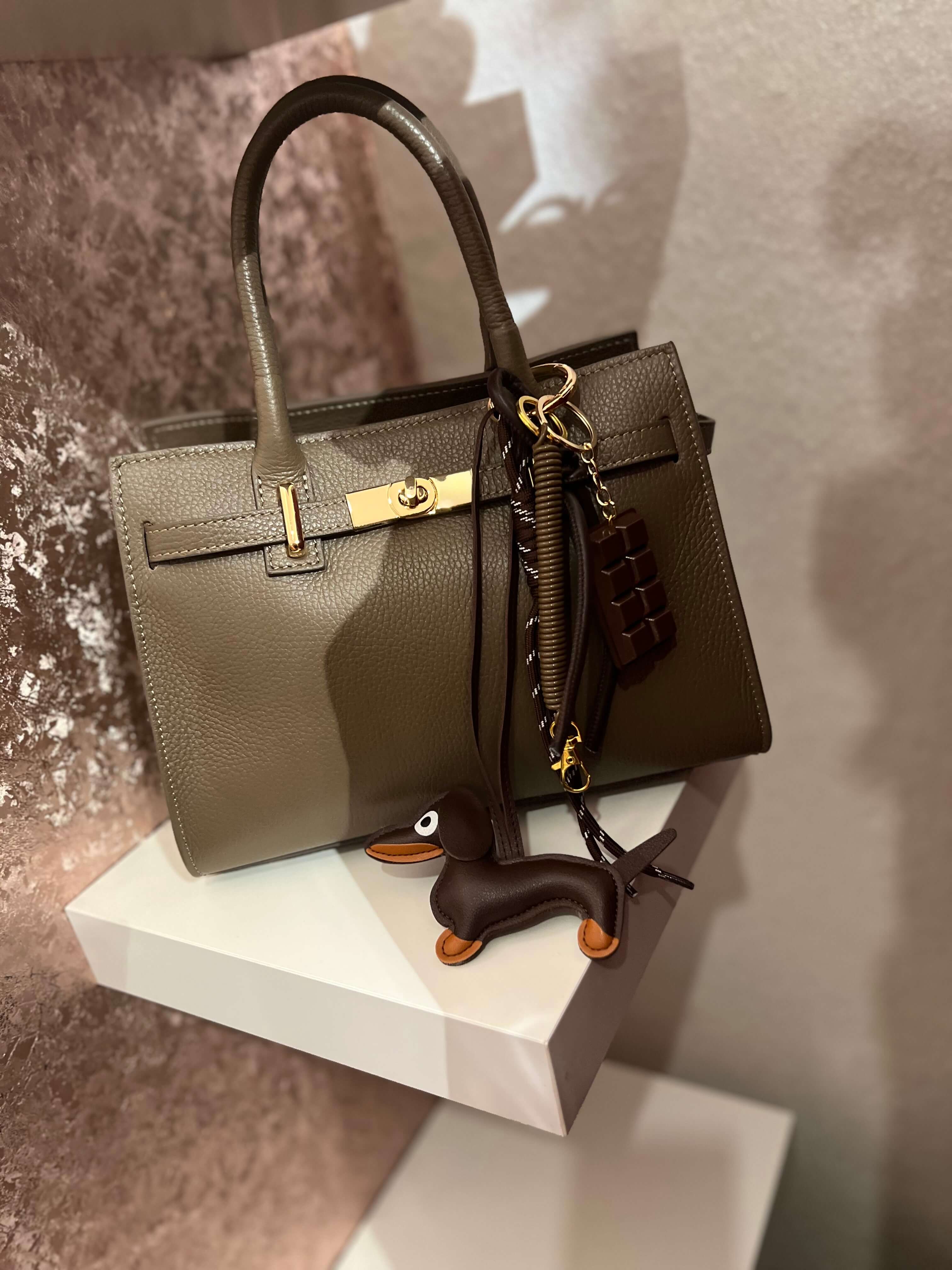 Taschenanhänger in Form eines braunen Dackels aus Kunstleder, befestigt an einer olivfarbenen Handtasche mit goldfarbenem Clip und zusätzlichen dekorativen Anhängern.