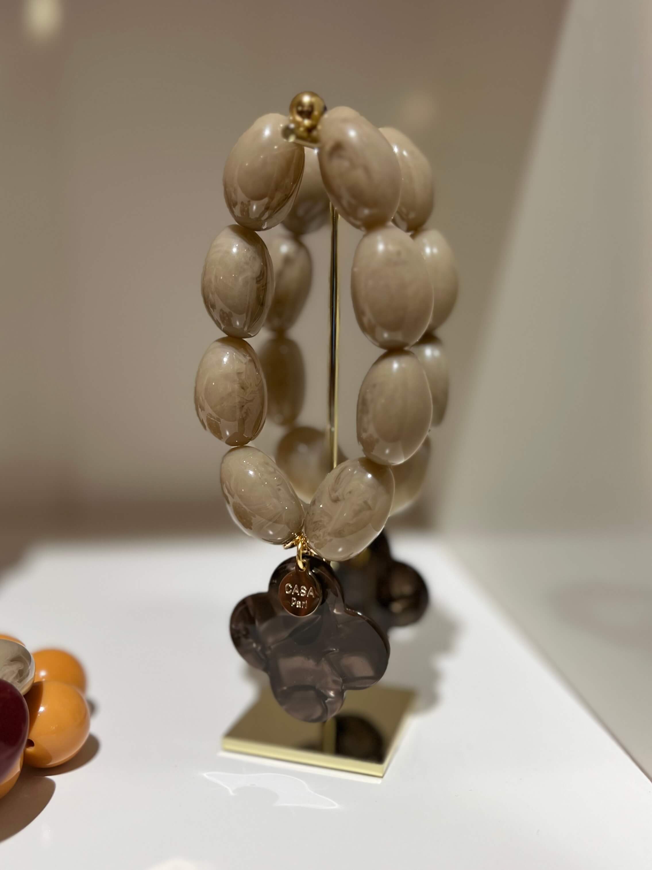 Armband aus beigen Kunstharzperlen mit dunkelbraunem Kleeblattanhänger, stehend auf einem goldfarbenen Ständer vor neutralem Hintergrund.