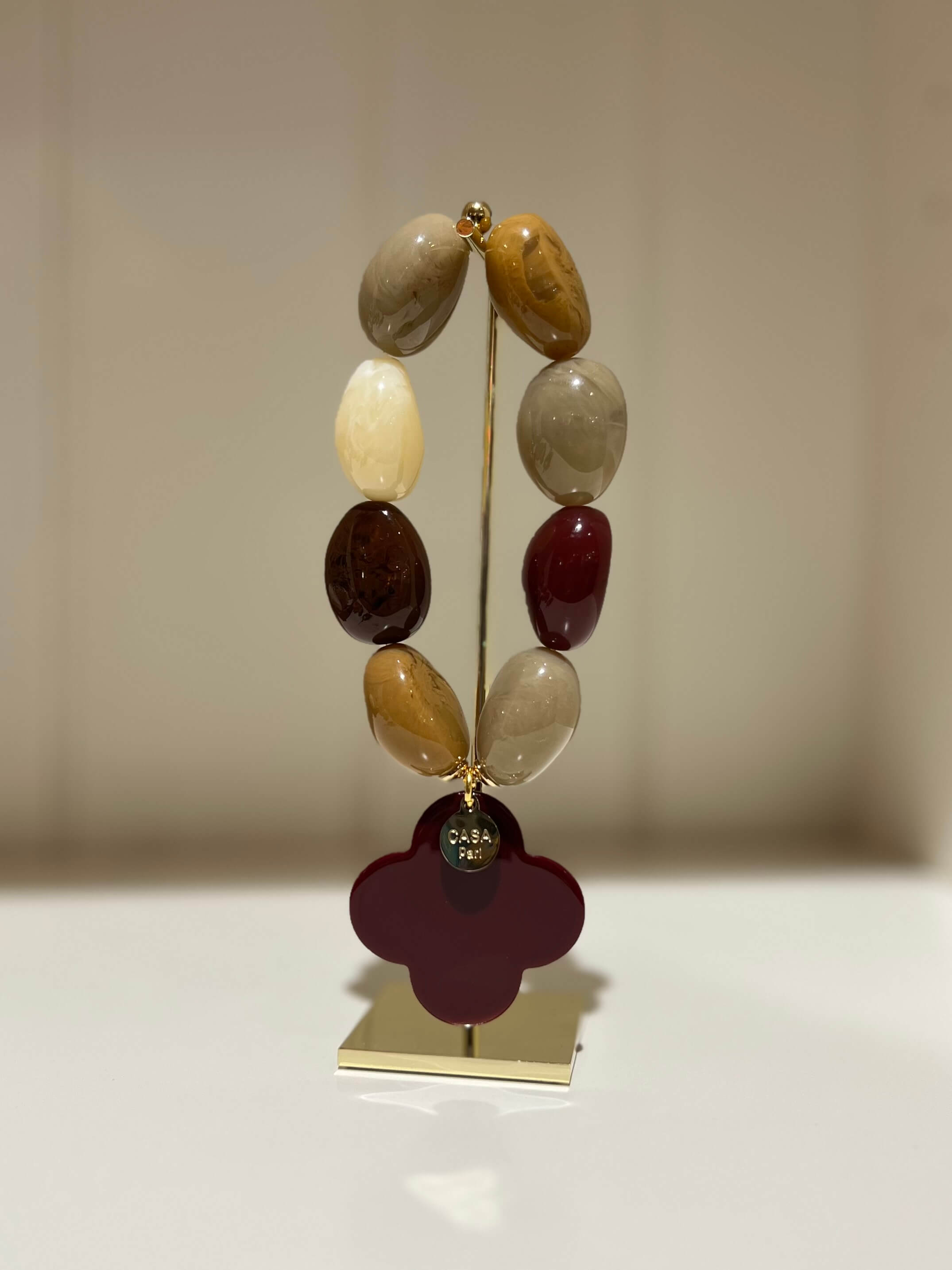 Armband aus bunten Kunstharzperlen mit dunkelbraunem Kleeblattanhänger, stehend auf einem goldfarbenen Ständer vor neutralem Hintergrund.