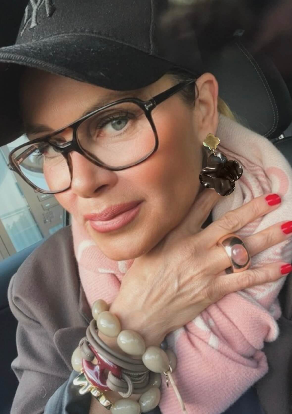 Person mit schwarzer Baseballkappe, Brille und dunklen Kleeblatt-Ohrring. Die Hand trägt mehrere Armbänder und einen Ring mit rundem Stein.
