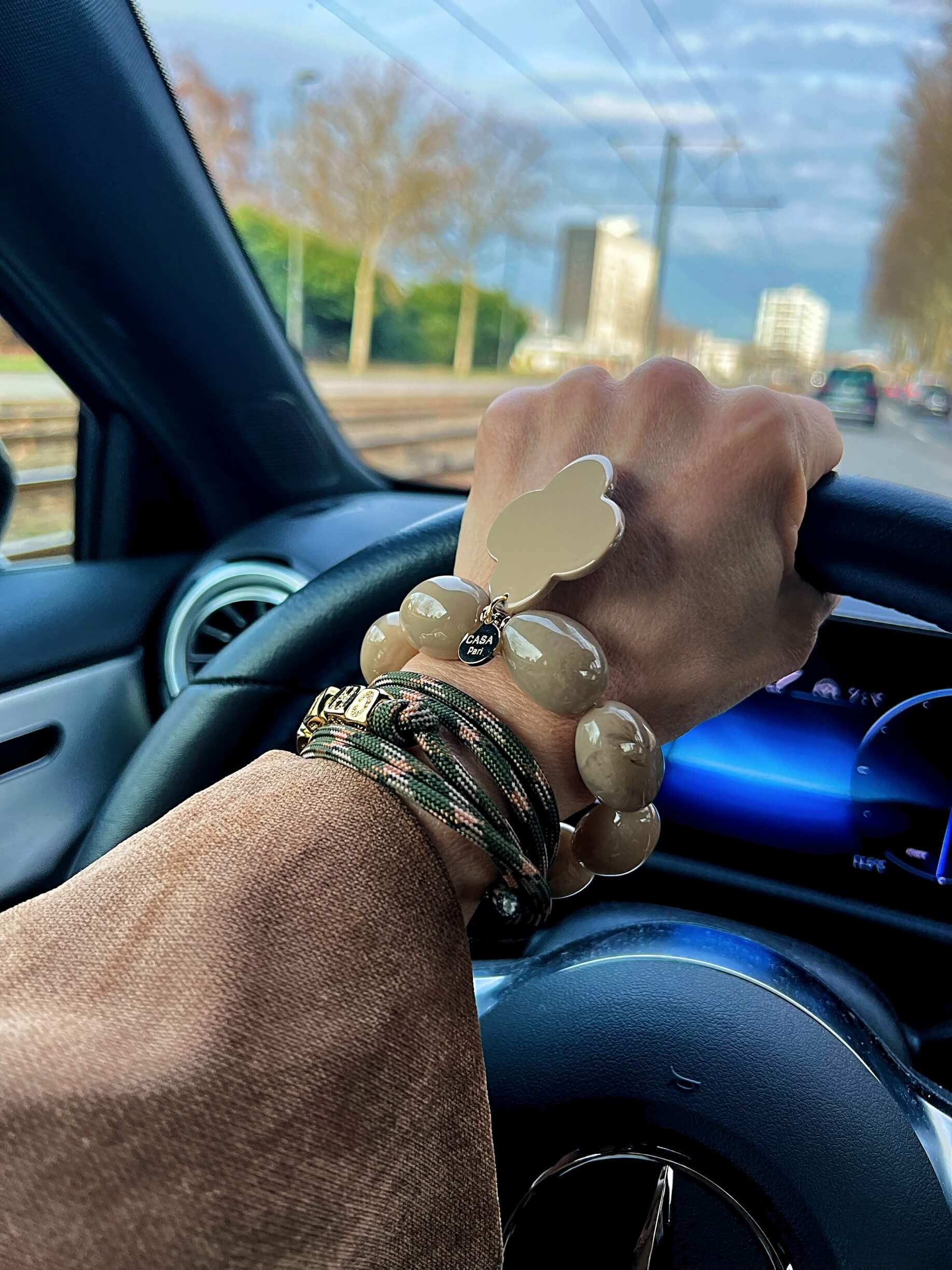Hand am Lenkrad eines Autos, getragen werden mehrere Armbänder in Natur- und Grüntönen, darunter ein Perlenarmband mit Kleeblatt-Anhänger und geflochtene Armbänder, aufgenommen im Alltag bei Tageslicht.